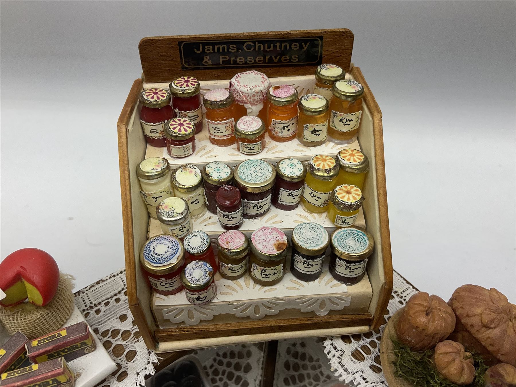Collection of miniature dolls house grocery shop furniture and produce, comprising glazed corner counter with brass style cash register, shelved preserves unit, pies, cheese board and trays of meats, pair of white cheese counters, biscuit display with six tins, glazed preserves cabinet, pair of shop chairs and four further cheese boards complete with grapes and label flags