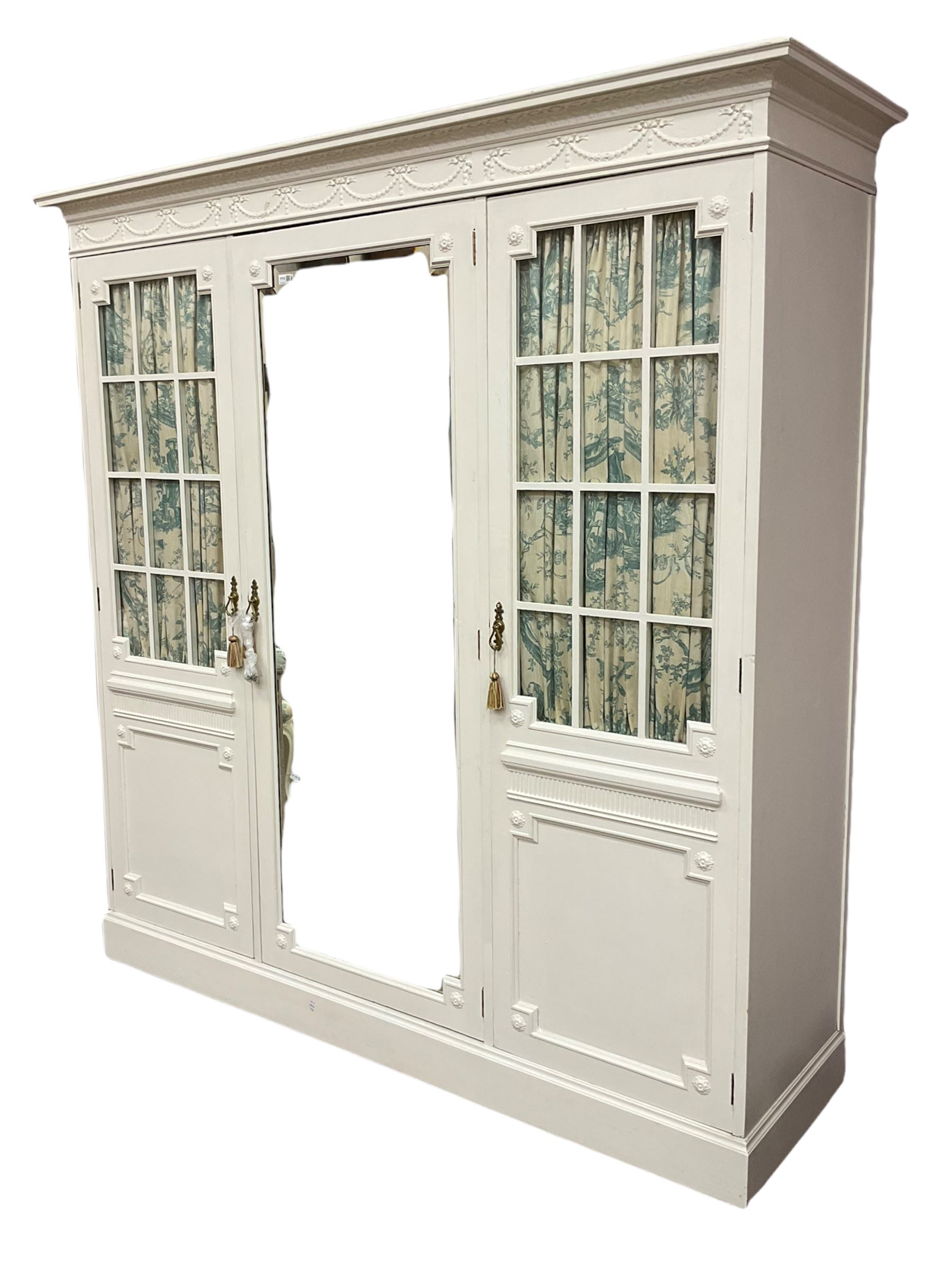 Maple and Co - Edwardian Hepplewhite design white painted triple wardrobe, foliate carved projecting cornice over ribbon and husk swag decorated frieze, two curtain lined astragal glazed doors and centre full mirror door, fitted mahogany interior including slides and drawers, plinth base, label to top of central door