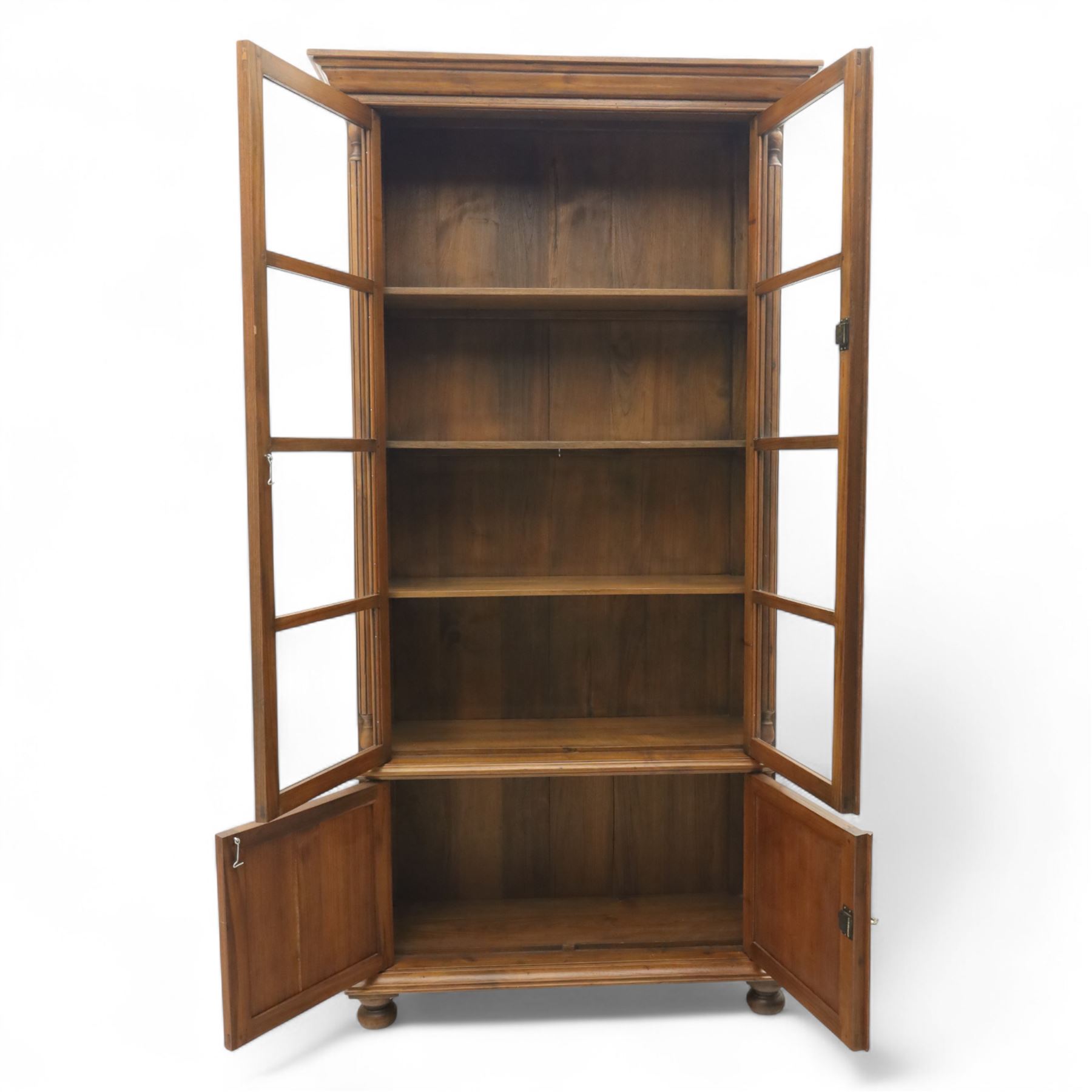 Pair of late 20th century Thai hardwood display cabinets, each with moulded cornice over twin glazed doors enclosing adjustable shelves, panelled cupboard doors below flanked by fluted columns, raised on bun feet