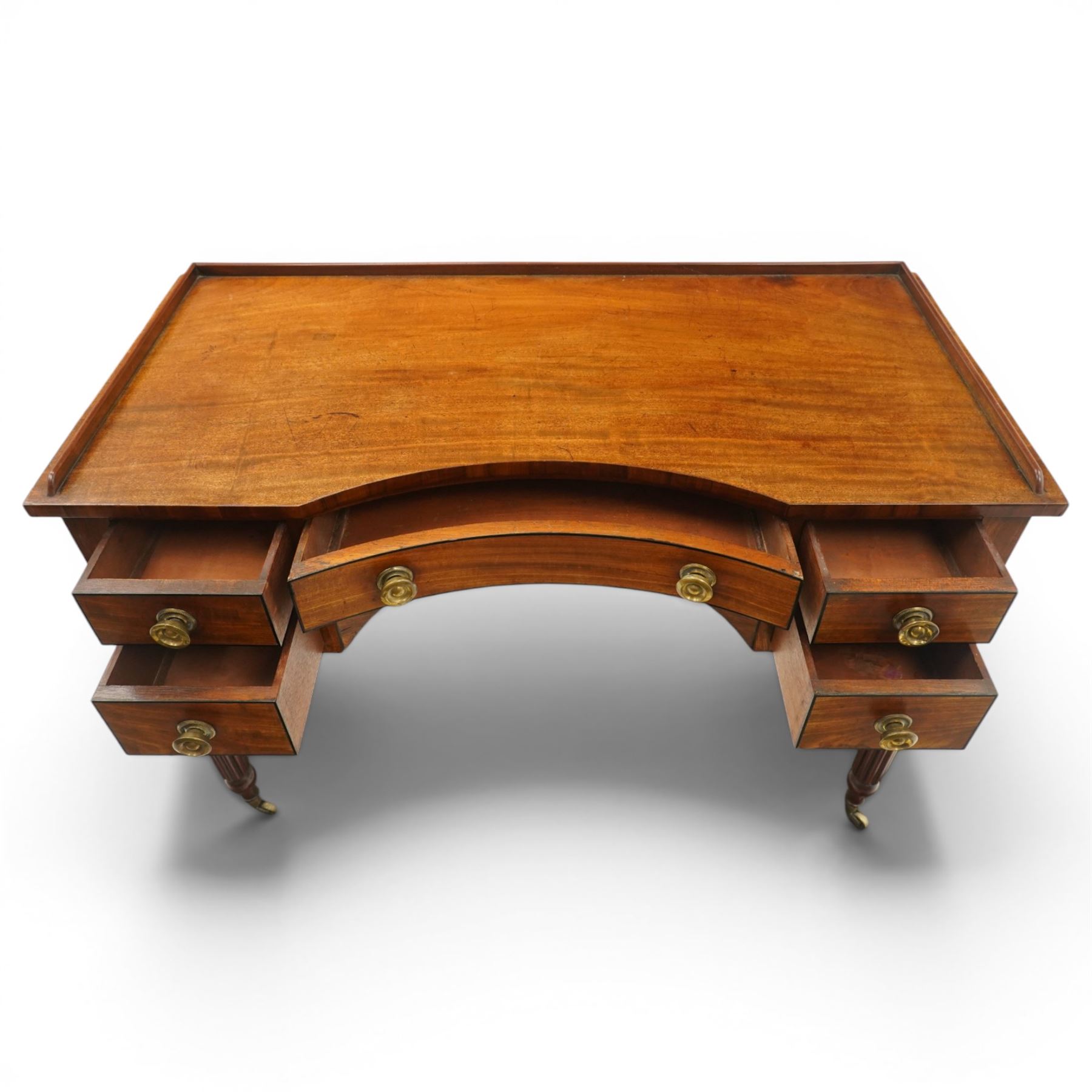In the manner of Gillows - Regency period mahogany kneehole dressing table or writing desk, the shaped rectangular top with raised three-quarter gallery, fitted with central concave frieze drawer flaked by two graduating drawers to each side, each with ebony stringing, raised on turned and reeded supports terminating to brass cups and castors