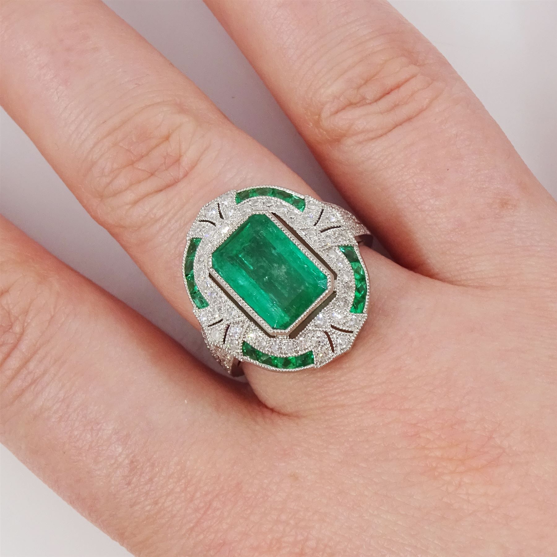 Platinum emerald and diamond milgrain set dress ring, single octagonal cut emerald of 2.40 carat, surrounded by single cut diamonds and a border of calibre cut emeralds, with fancy pierced design gallery diamond and engraved flower head shoulders, stamped 850 Plat