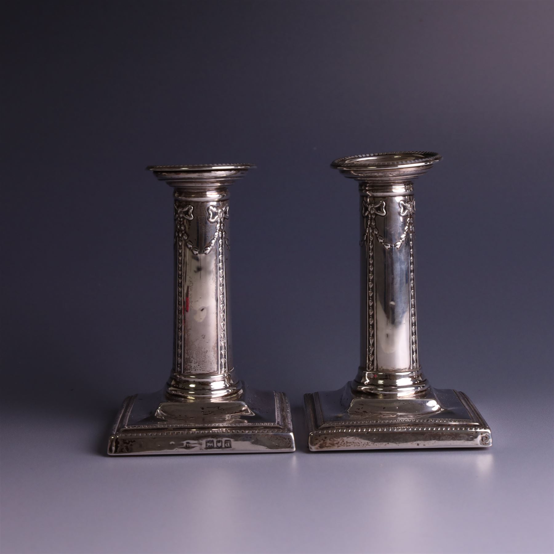 Pair of late Victorian silver mounted candlesticks, the stems embossed with classical ribbon and swag design, upon weighted stepped square bases, hallmarked London 1899, maker's mark worn and indistinct, H12cm
