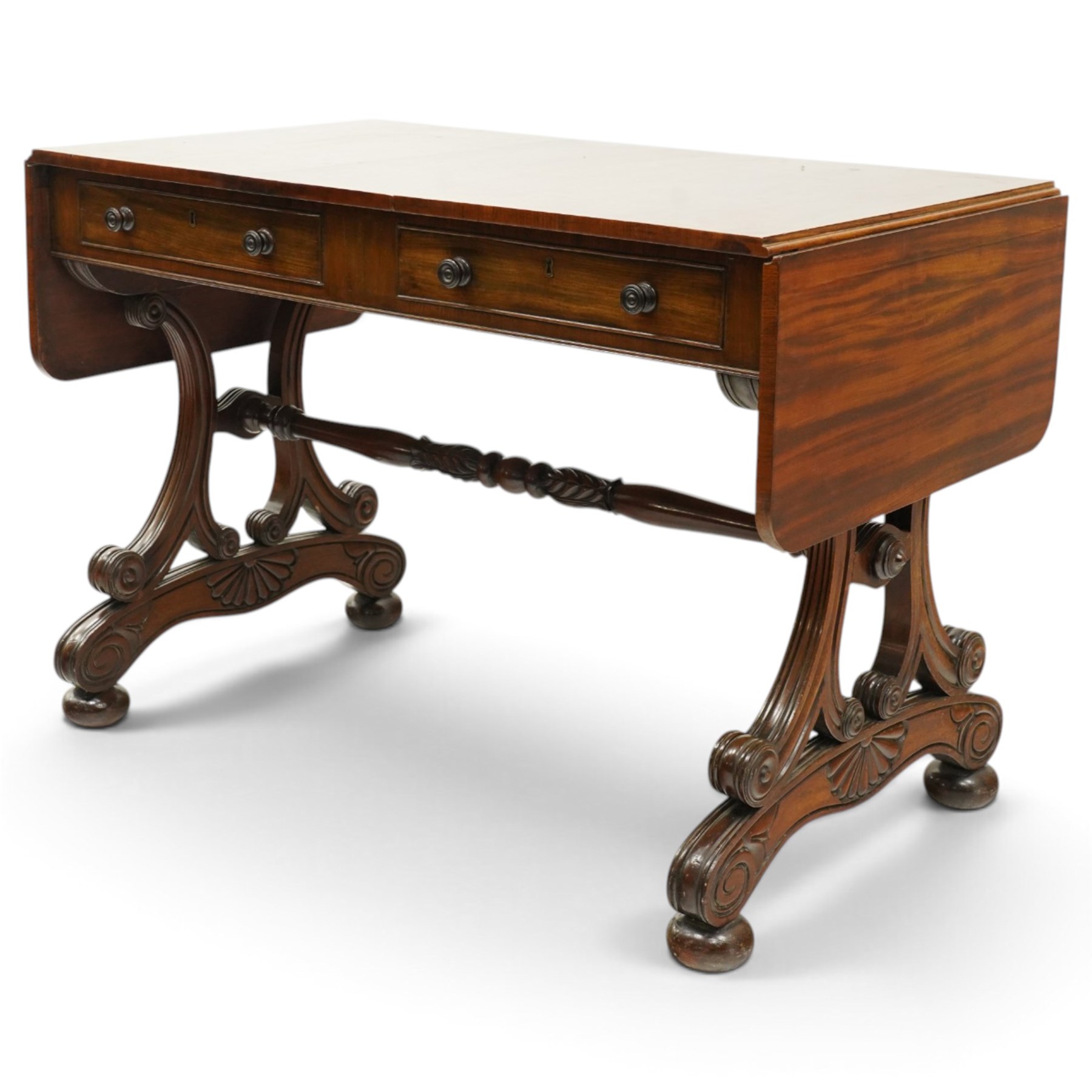 Fine 19th century figured mahogany sofa table, the rectangular twin drop-leaf top with rounded corners above two cock-beaded frieze drawers with opposing faux drawers, fitted with turned handles, raised on twin reeded and C-scroll-carved end supports with palmette-carved platform bases and compressed bun feet, united by a turned and foliate-carved stretcher