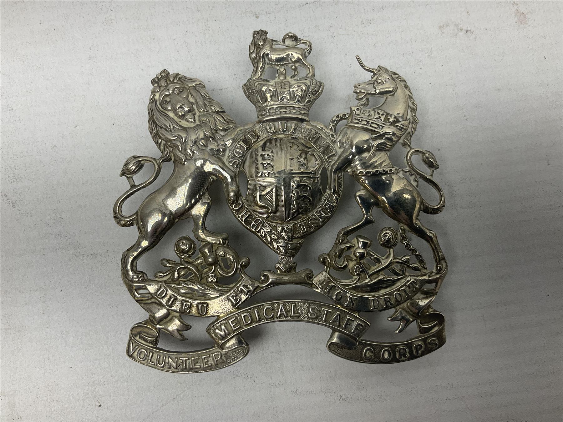 Victorian Volunteer Medical Staff Corps white metal helmet plate in the form of Victorian crowned Royal Arms with three part lower scroll for “VOLUNTEER MEDICAL STAFF CORPS”; three lug fittings to the reverse L8.5cm