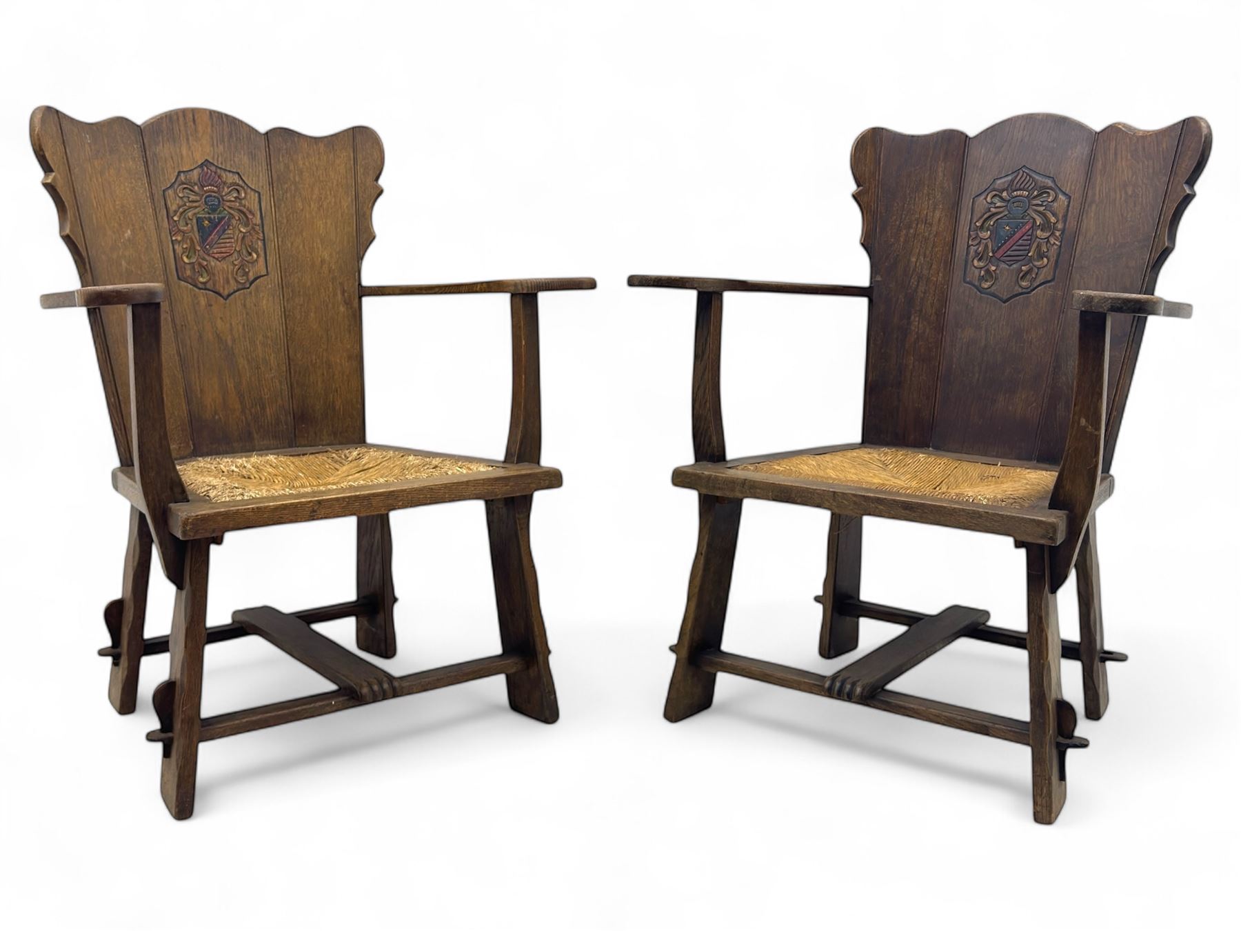 Pair of Arts and Crafts oak armchairs each with shaped backrest centred by polychrome painted crest, rush seat over straight square legs united by shaped central stretcher