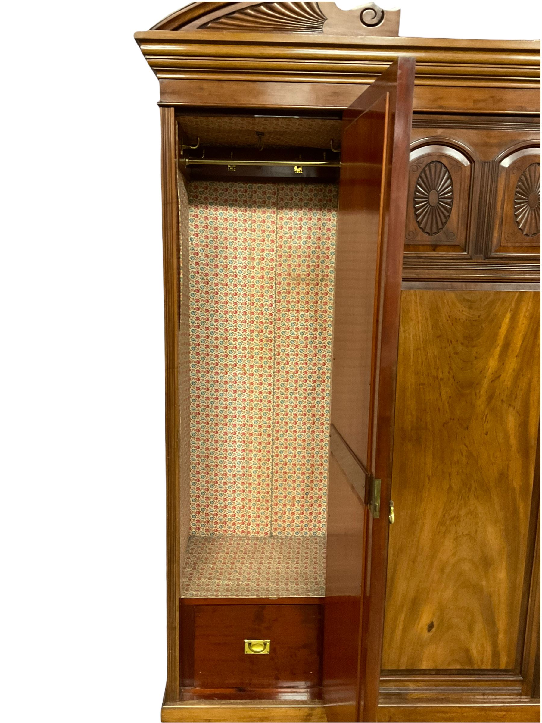 Victorian mahogany triple wardrobe, the pediment relief carved with urn and linen swags, the central panelled door carved with two fan motifs enclosing slides and drawers, two mirror glazed doors with bevelled plates enclosing hangings space and drawers, plinth base