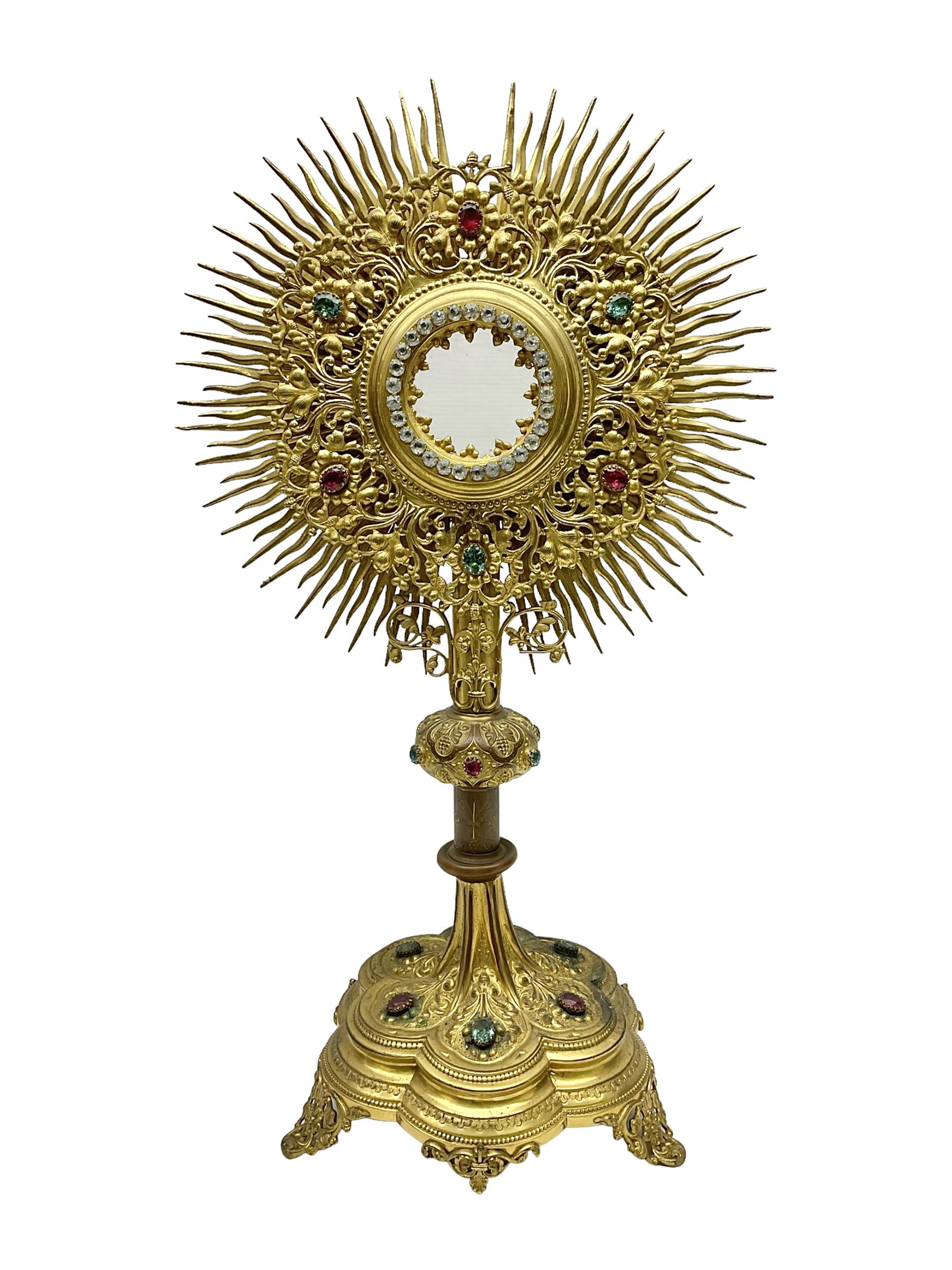 20th century gilt metal monstrance, in the Gothic revival style, with glazed aperture to centre surrounded with clear paste stones, in a pierced floral and scroll border set with coloured paste stones, within a sunburst surround, on a paste set knopped stem and similar foliate foot, H59.3cm