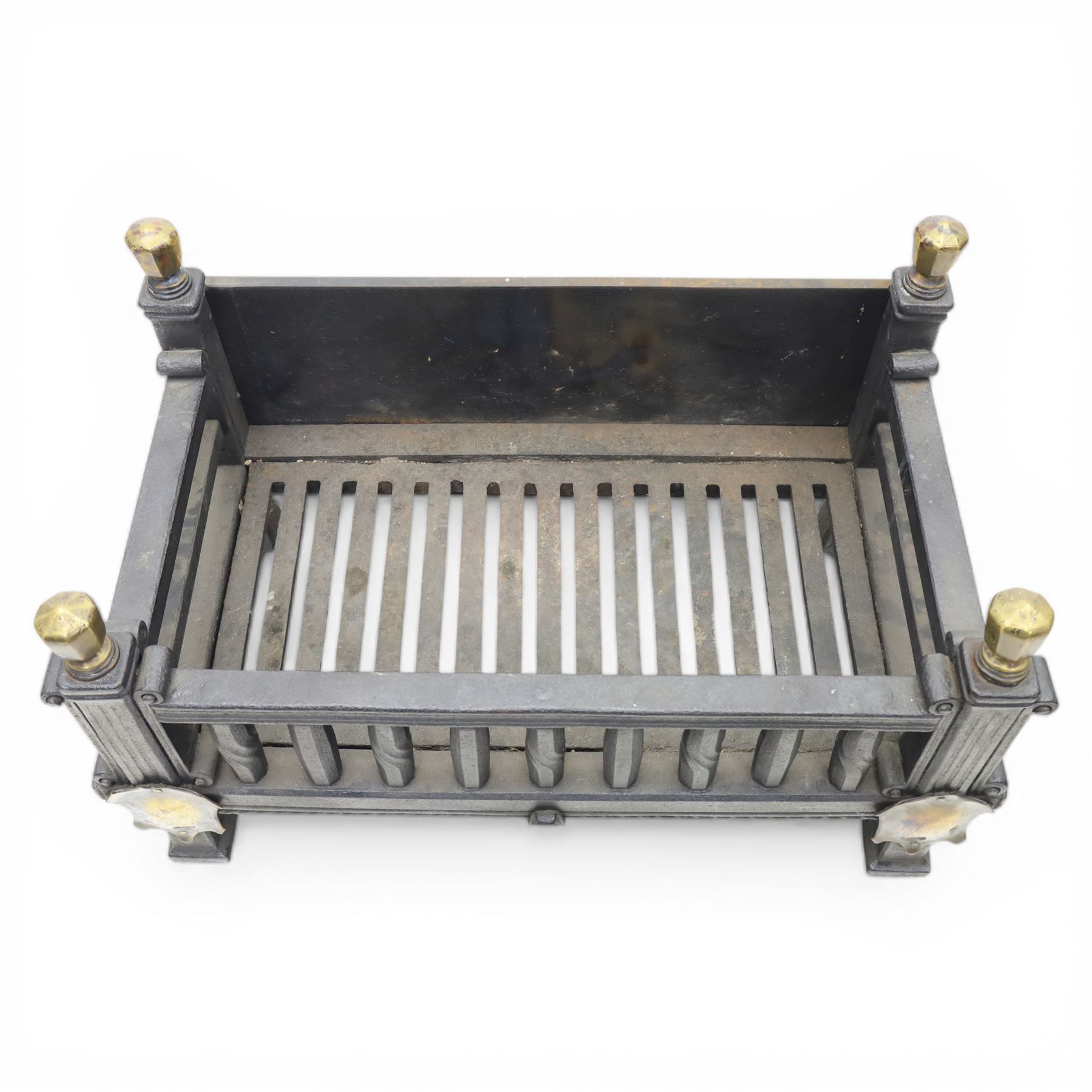 Arts and Crafts cast iron fire grate, rectangular backplate with plain upper rail over a slatted front of cylindrical tapering bars, flanked by square section uprights with fluted detail and scroll terminals, raised on stepped feet with shaped brass roundel mounts and brass finials to the corner posts