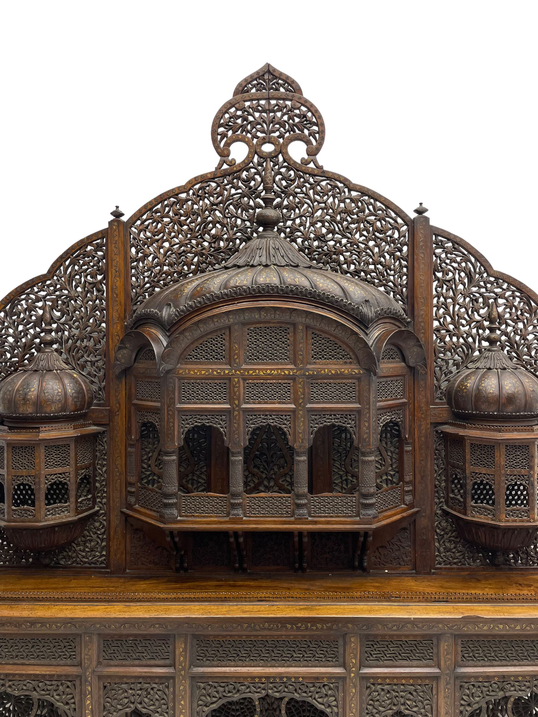 Late 19th century Indian Burmese carved and pierced hardwood and brass inlaid wall shelf, the shaped back profusely decorated with trailing and interlaced foliage, with projecting architectural structures, the central balcony with canopy top and five open windows with arched apertures on columns, two flanking five-sided balconies, the shelf supported by six foliate carved columns with pierced cusped pointed arches 