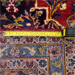 Central Persian Kashan hand-knotted carpet, crimson central field with lobbed medallion in navy blue and ivory ground, surrounded by a dense all-over floral design of scrolling vines, palmettes, and stylized blossoms, enclosed within a deep navy main border, flanked by multiple guard stripes with fringe ends 


