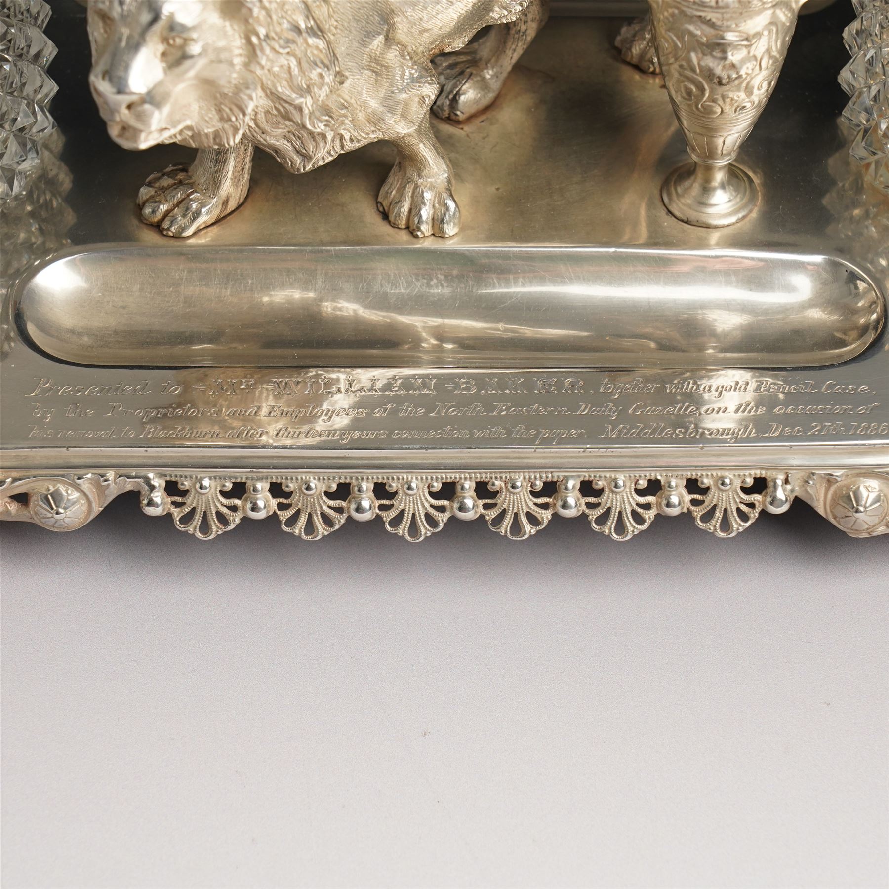 19th century Walker & Hall, silver plated inkstand, oblong and on four paw feet, the border decorated with pierced shell design, each side with a pen-rest, with two cut-glass bottles and central lion and urn finials, engraved to the front with an inscription, H15cm, L30cm