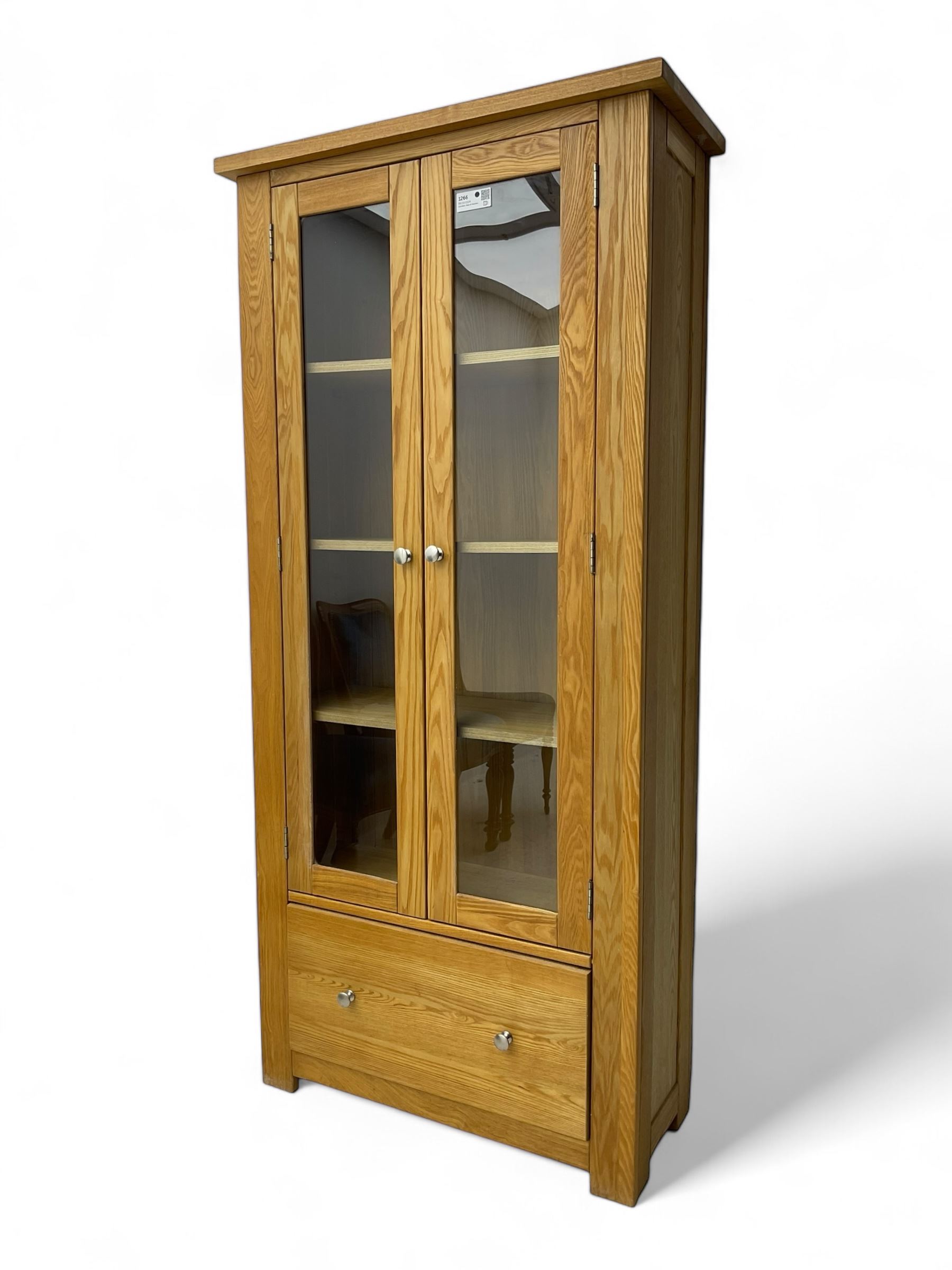 Light oak bookcase display cabinet, projecting square cornice over glazed double doors enclosing three adjustable shelves, single drawer base, on stile supports