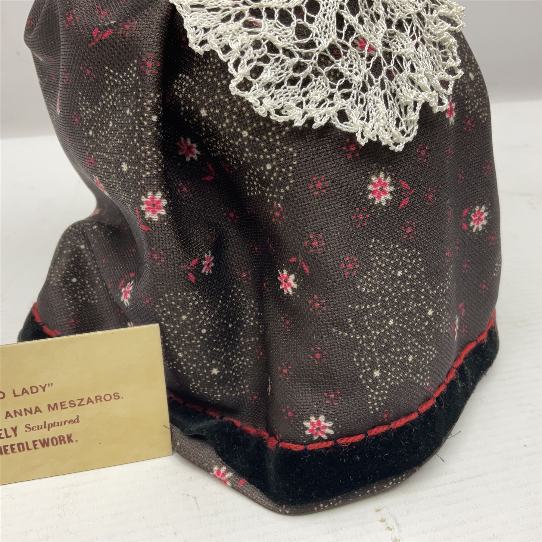Anna Meszaros Hungary - hand made needlework figurine 'Old Lady' seated on a stool wearing a lace trimmed black/white/red floral full length dress and hat, working on a piece of lace H30cm  Auctioneer's Note: Anna Meszaros came to England from her native Hungary in 1959 to marry an English businessman she met while demonstrating her art at the 1958 Brussels Exhibition. Shortly before she left for England she was awarded the title of Folk Artist Master by the Hungarian Government. Anna was a gifted painter of mainly portraits and sculptress before starting to make her figurines which are completely hand made and unique, each with a character and expression of its own. The hands, feet and face are sculptured by layering the material and pulling the features into place with needle and thread. She died in Hull in 1998.