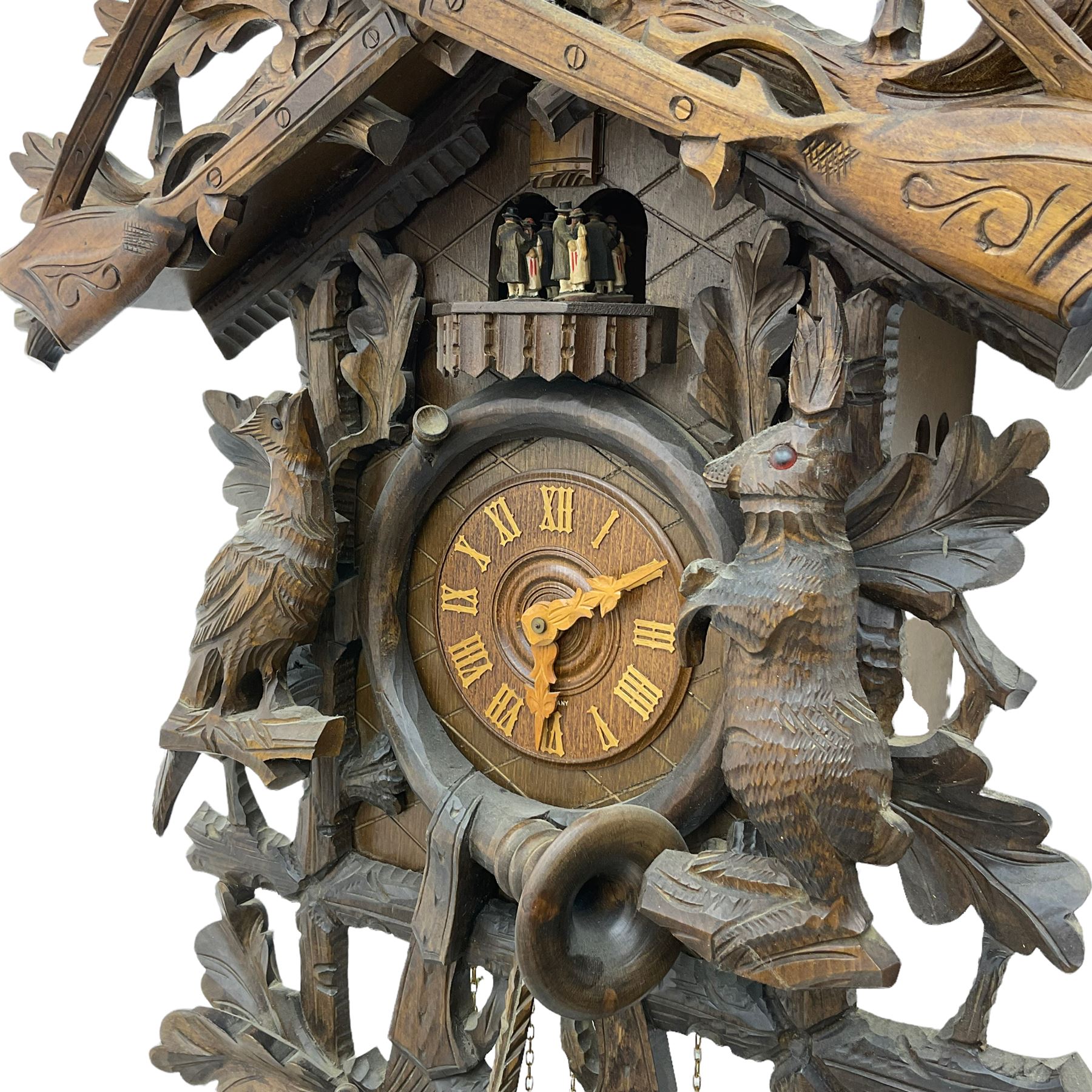A large and imposing 20th century three train 30hr cuckoo clock with automata and musical features, profusely carved case with a carousel, animals, leaves and sporting guns, dial with pierced wooden numerals and wooden hands, with three weights and pendulum, strike/silent facility. 
