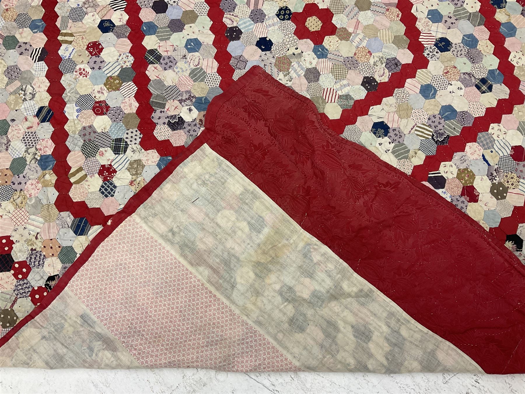 19th century patchwork quilt, hand stitched from hexagons of various materials including printed patterned and floral examples, featuring repeating large central hexagon to centre, the top and bottom with thick panel red jacquard border, the sides bordered with plain red fabric, lined with panels to reverse, 180 x 190 cm