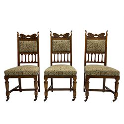 Set six Edwardian oak dining chairs, the cresting rails relief carved with foliate scrolls and pierced with handle, upholstered in foliate pattern fabric decorated with heraldic shields,. turned front supports on castors joined by plain H-framed stretchers
