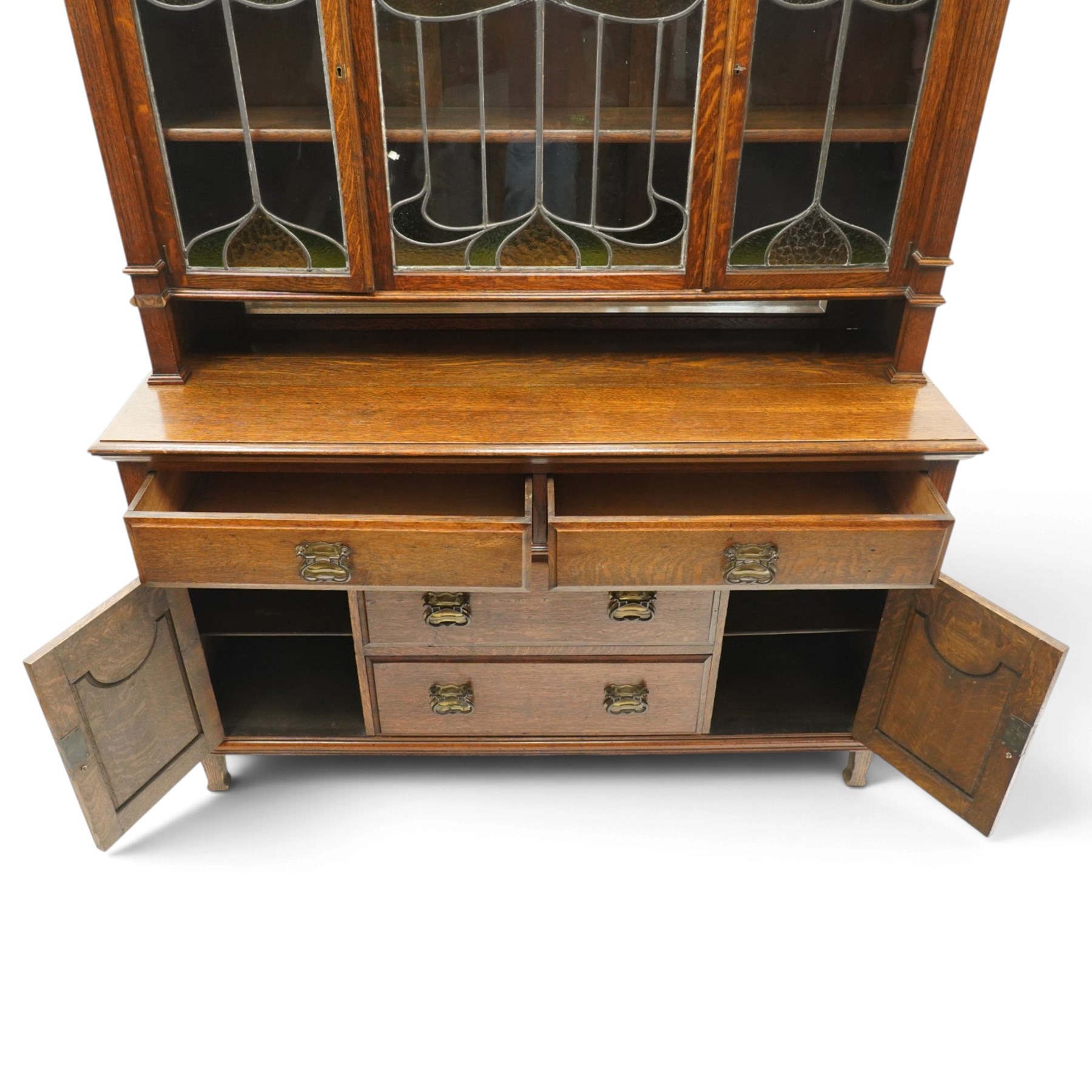 Arts and Crafts oak dresser, projecting moulded cornice above three glazed panels with coloured leaded glass floral panels, flanked by two glazed doors enclosing two adjustable shelves, open mirrored recess below with mirrored back, the base fitted with two short drawers over two central drawers flanked by carved panelled cupboard doors, ornate brass handles, raised on tapering square legs