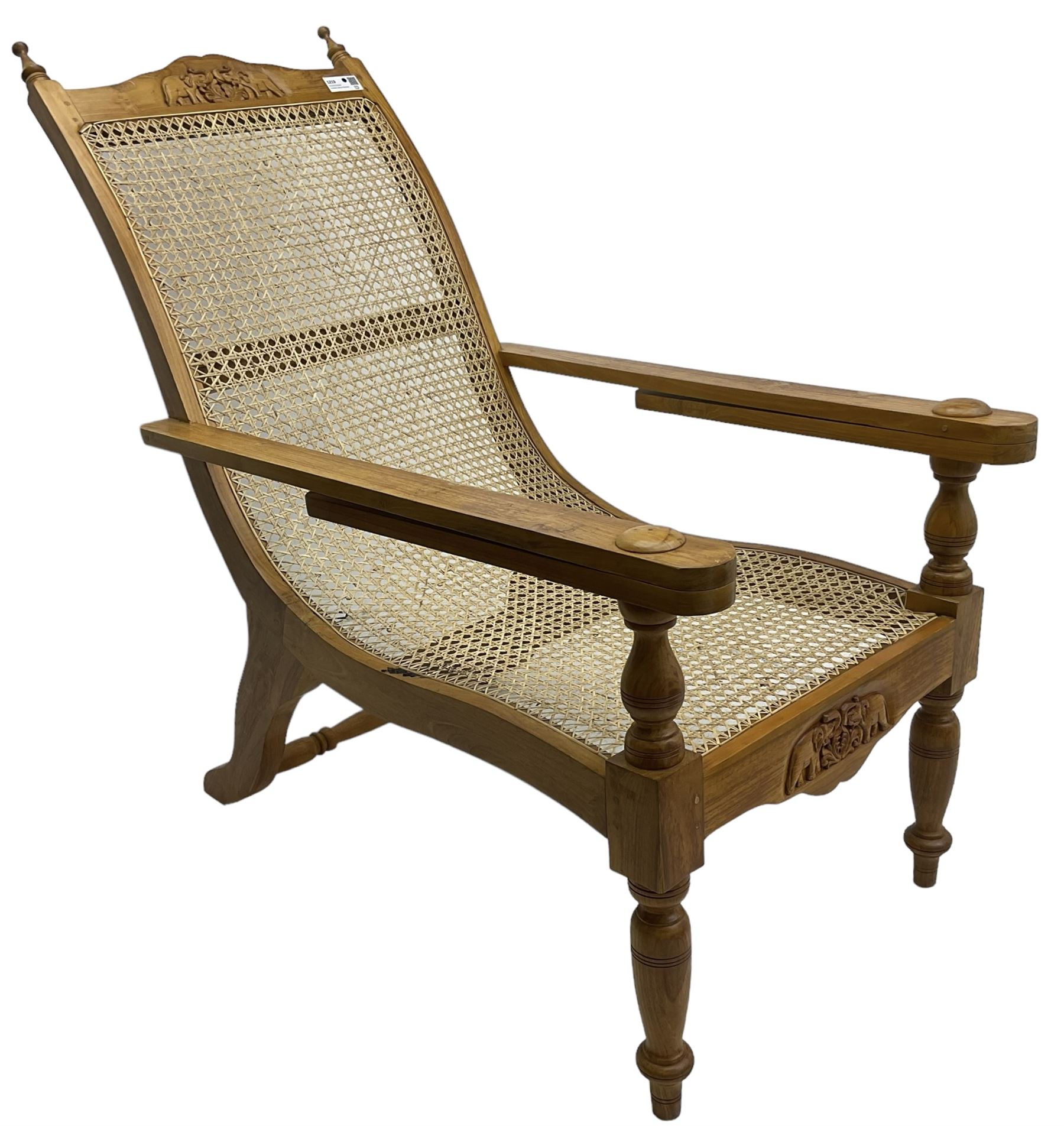 Late 20th century teak framed plantation chair, cresting rail and apron carved with two elephants with upraised trunks, flanked by finials, cane back and seat, fitted with hinged extending leg rests, on ring turned supports