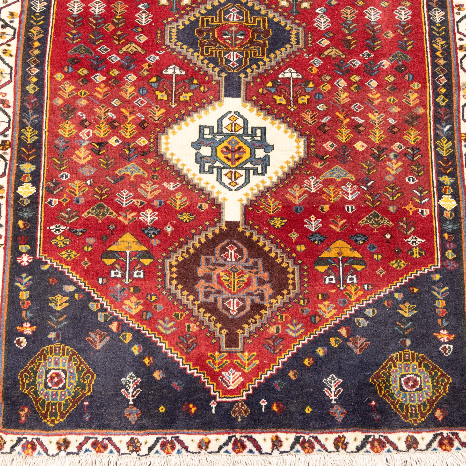 South West Persian hand-knotted Qashgai runner, madder-red field with a central column of linked diamond medallions in tones of indigo, ochre, ivory, and green, surrounded by stylized floral and boteh motifs within a cream border of angular vines and serrated devices, with cotton fringe ends   
