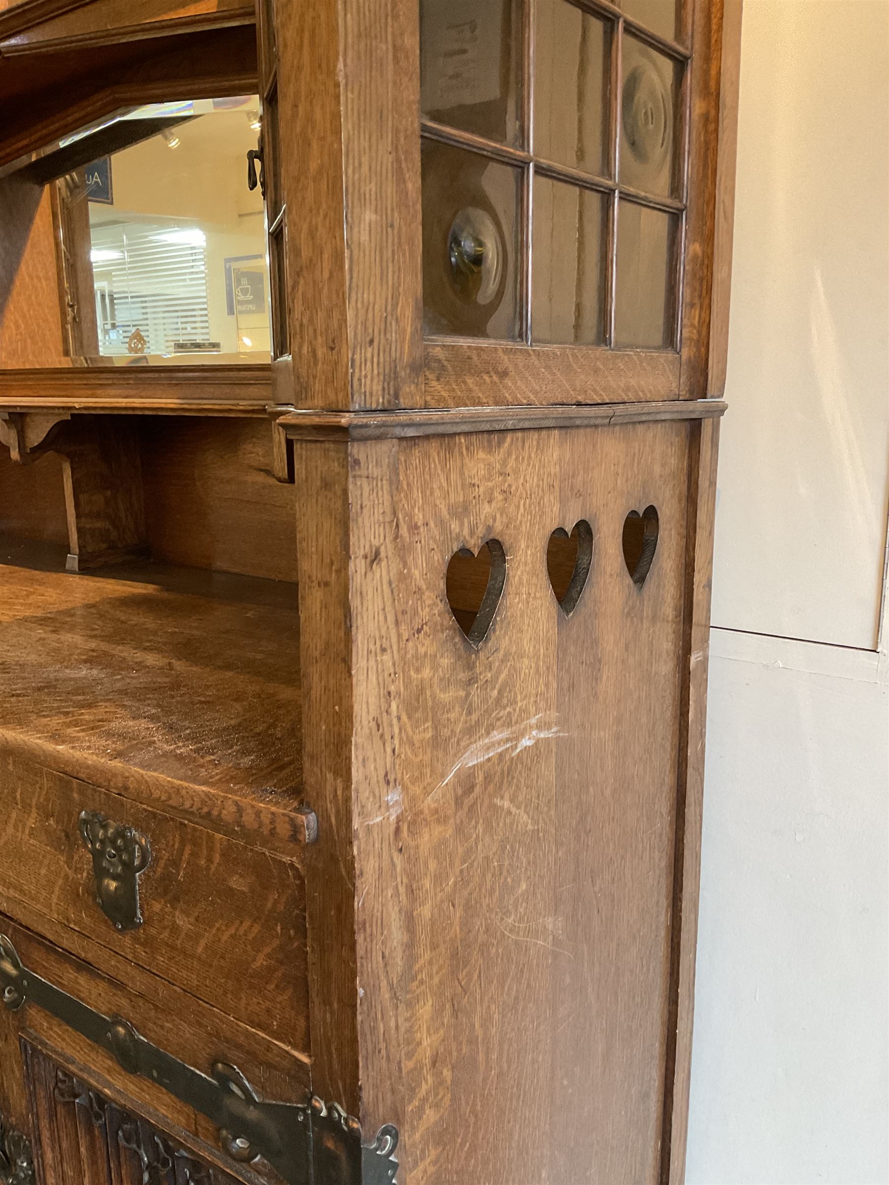 Shapland and Petter - Arts & Crafts period oak dresser, raised gallery top with spade balustrade, central bevelled mirror flanked by two cabinet, astragal glazed doors with yellow tinted glass and bullseye panes, the sides with pierced heart shaped decoration, fitted with two drawers and two double cupboards, enclosed by linenfold panelled doors,  copper work strap hinges and fixtures, on shaped spade feet, the locks stamped 'S&P'
