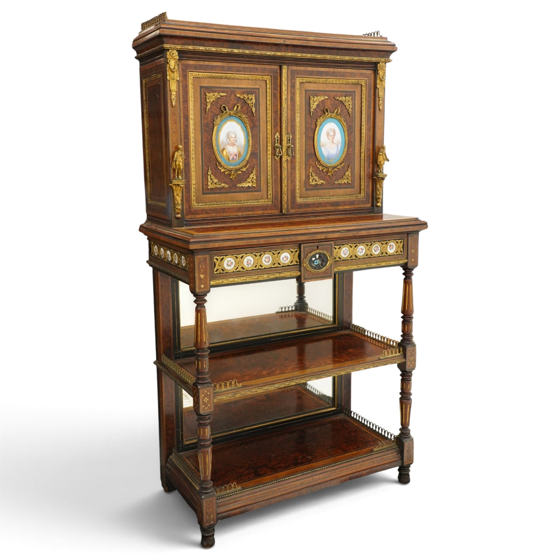 Fine late 19th century Aesthetic Movement ebony and amboyna cabinet on stand with parcel-gilt detail, the moulded cornice with pierced gilt-brass three-quarter gallery above a pair of panelled doors centred by oval porcelain portrait plaques within gilt-metal ribbon-tied frames, the doors further enriched with gilt-metal foliate mounts and flanked by classical figural terms, enclosing a shelved interior, the projecting lower section with amboyna top above a frieze drawer set with a series of floral porcelain roundels within pierced gilt-metal mounts and a central oval floral plaque, raised on turned and fluted supports with gilt-heightened detail, the mirrored back behind two amboyna shelves with pierced gilt-brass galleries, on turned feet
