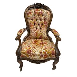 Victorian walnut framed open armchair, the pediment carved with flower heads and foliage, upholstered in buttoned floral fabric, shaped and carved arm uprights with scrolled terminals, on cabriole supports with brass castors