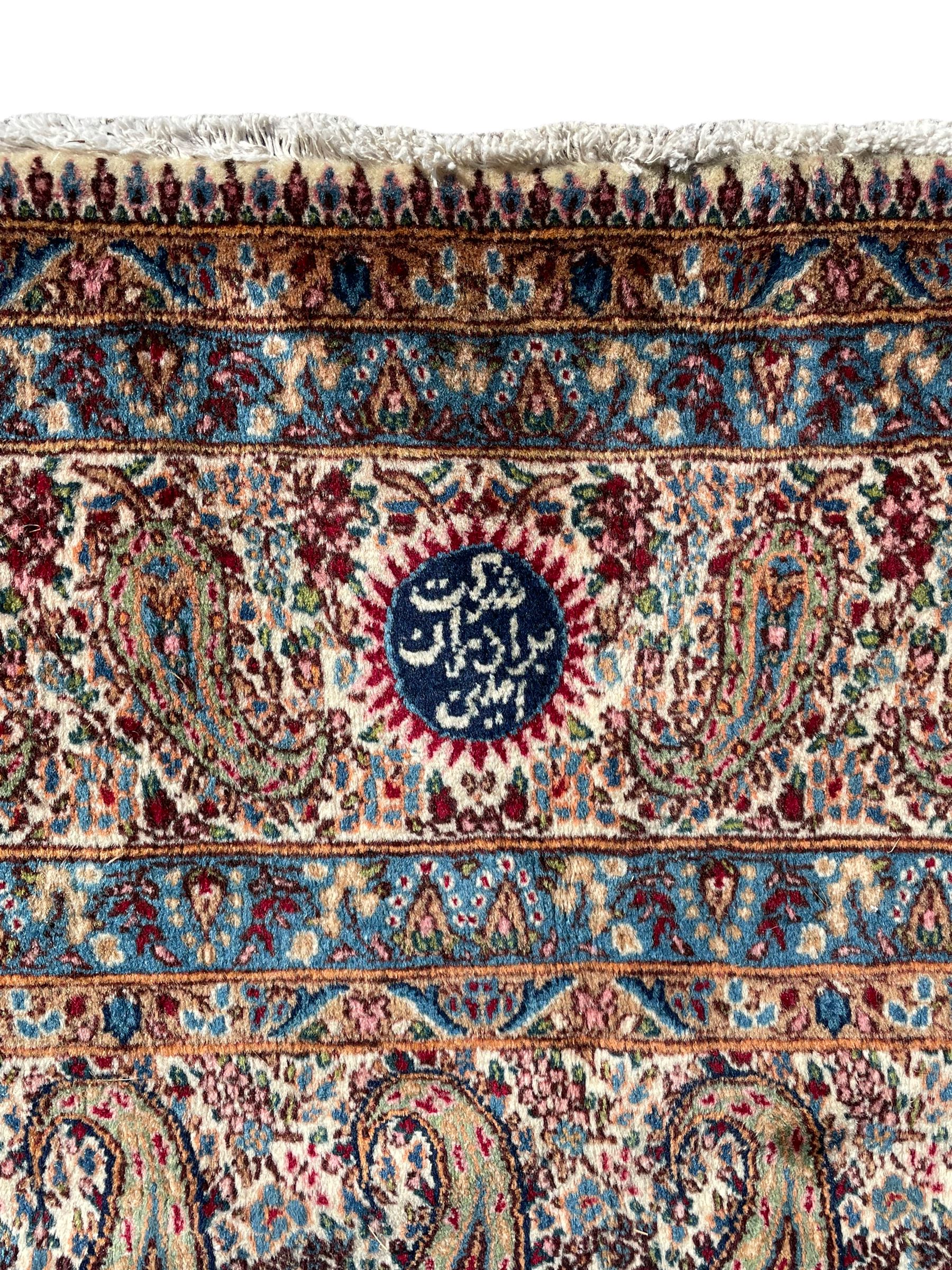 Persian Kirman carpet, the ivory ground field and border decorated with repeating Boteh motifs, interspaced with all-over floral design, blue ground border guards, the top border with signature panel