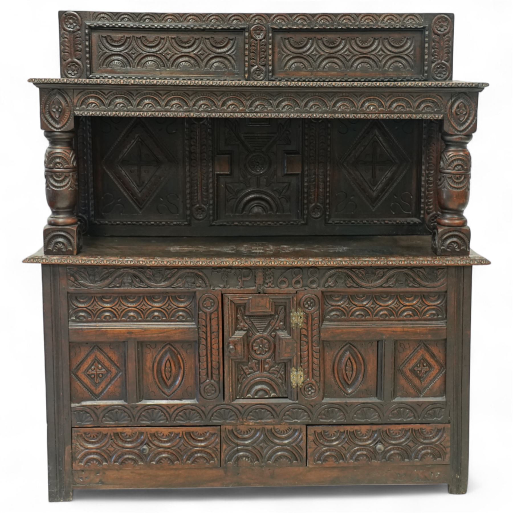 An early 20th-century Jacobean revival style carved oak Court Cupboard, tier-form of 17th-century design, rectangular form with carved back with geometric motifs and arches, projecting top section over twin baluster columns connecting the lower tier above a carved panelled front and panelled cupboard door with profusely carved lozenges and lunettes, above two base drawers, raised on block feet 