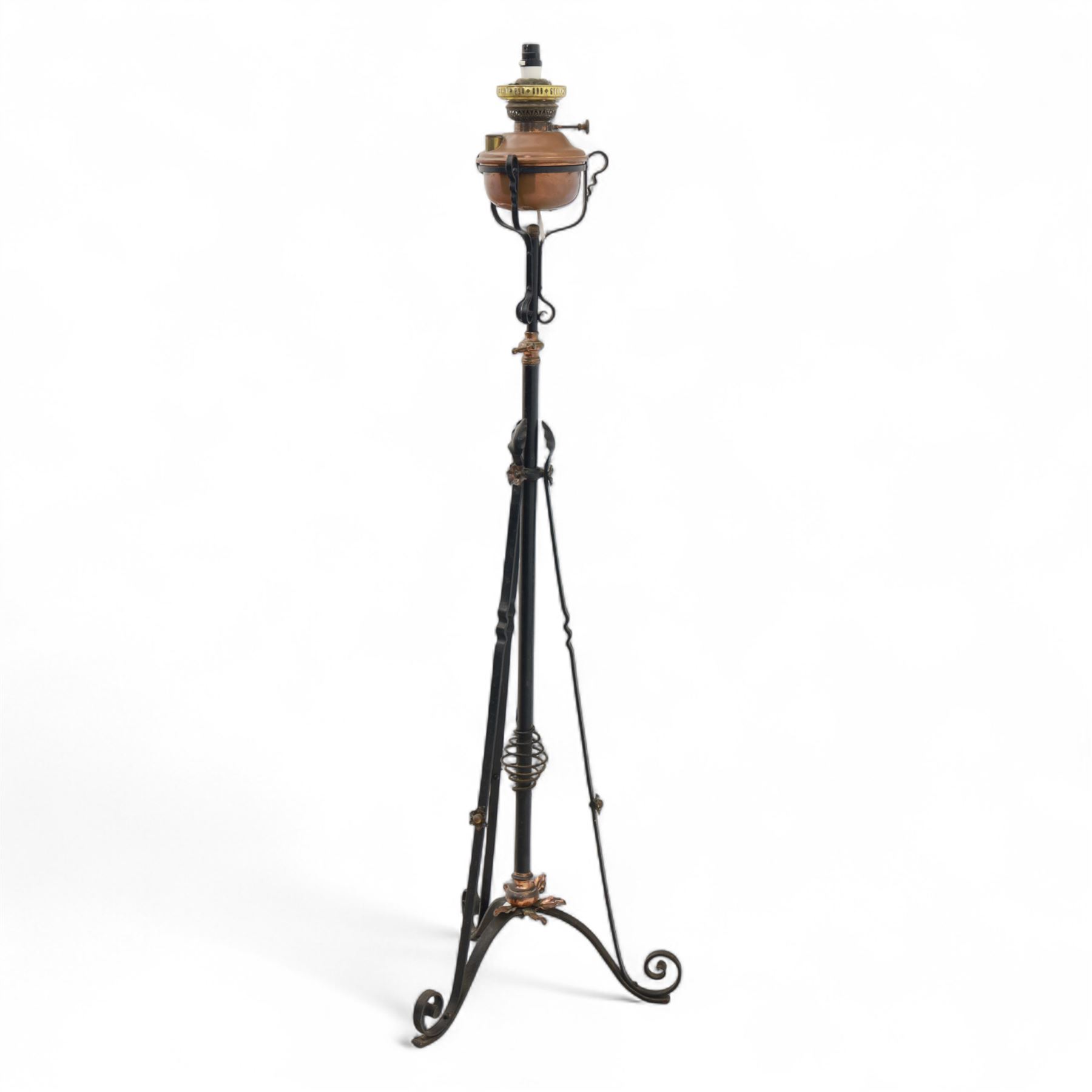 Early 20th century converted oil lamp standard, with copper reservoir and brass burner mount, later wired for electricity, on wrought iron tripod stand with scroll and twist ornamentation