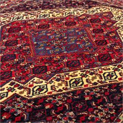 North West Persian Senneh hand-knotted rug, dark indigo field with a central bold stepped lozenge medallion of madder red and deep blue, enclosed within an ivory serrated contour forming a cruciform outline, medallion with repeating boteh and floral devices, field decorated with an all-over Herati pattern of rosettes enclosed by angular leaves principal border of madder red ground decorated with alternating floral heads, flanked by narrow guard stripes, with cotton fringe ends 