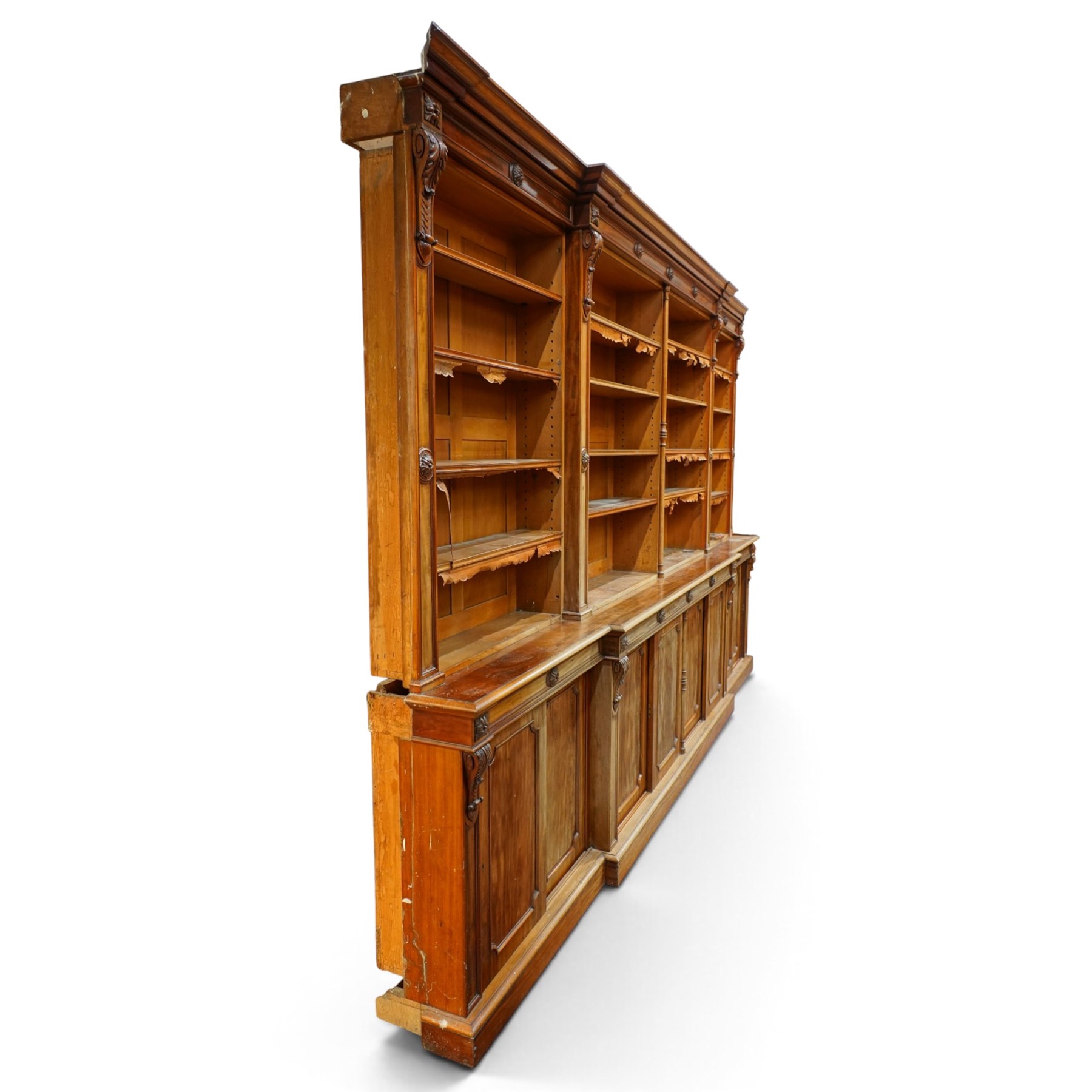 Large early 19th century Country House mahogany breakfront library bookcase, the projecting moulded cornice over a frieze applied with carved flowerhead paterae, the open panelled upper section fitted with adjustable shelves and shaped valances, divided by moulded pilasters with acanthus-carved corbels, the projecting base enclosed by eight panelled cupboard doors with central turned canted pilaster, on plinth base