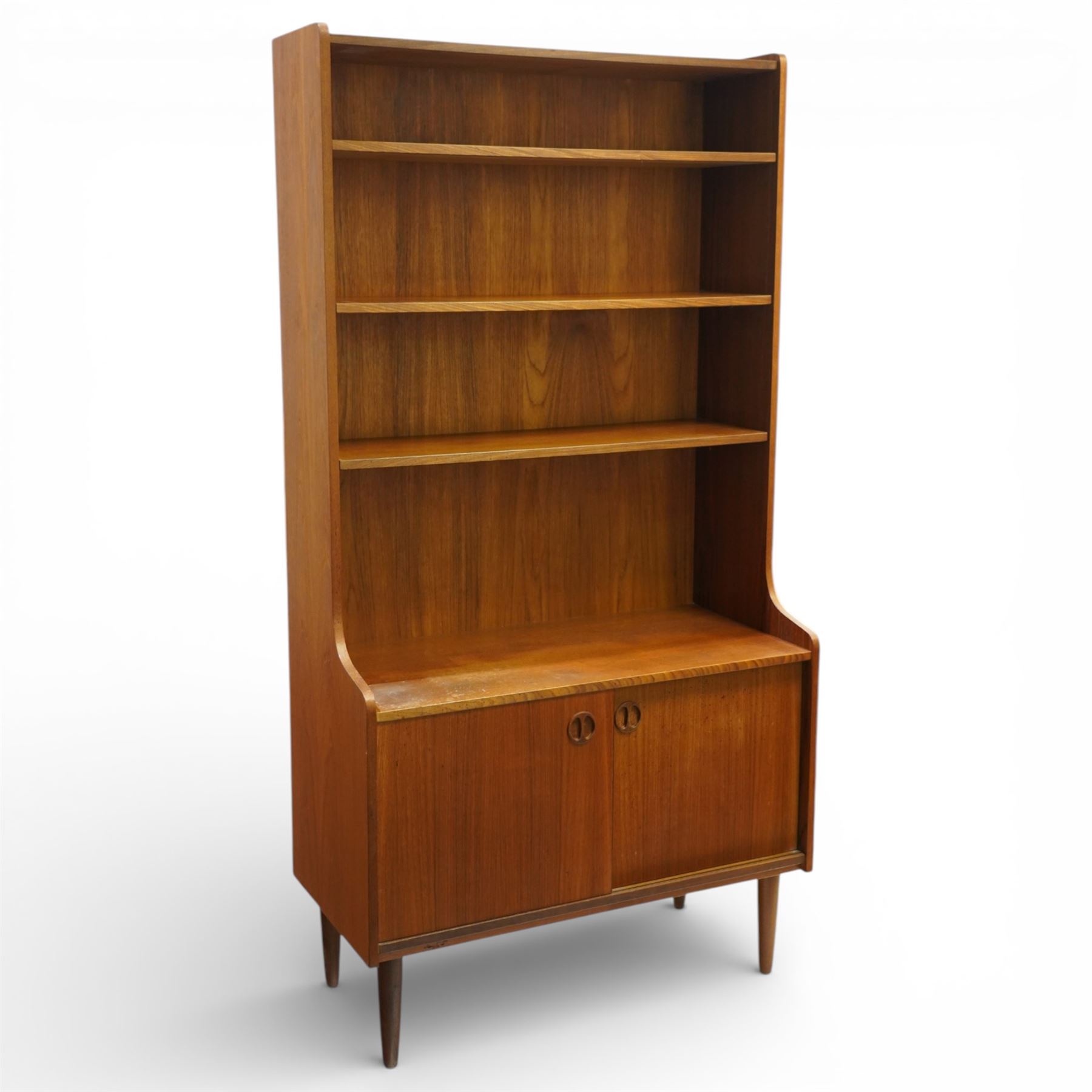 Mid 20th century Scandinavian teak bookcase cabinet in the manner of Johannes Sorth, the upper section with three open shelves over a shaped fall to a lower cupboard enclosed by sliding doors with recessed circular handles, raised on tapering supports