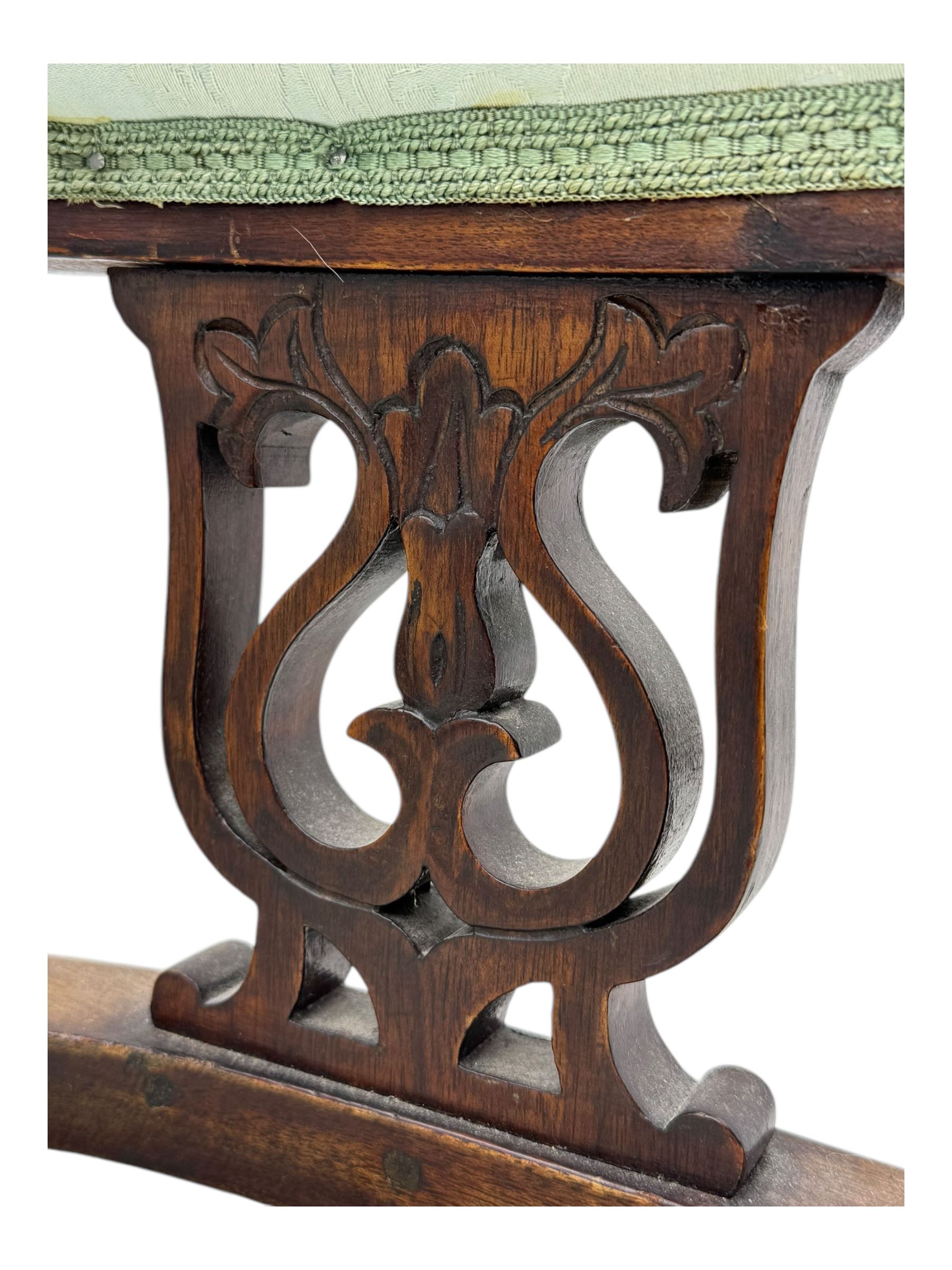 Late Victorian carved mahogany open armchair, the shaped back with fluted upholstered panel and padded arms above pierced scroll carved side supports, over a broad upholstered seat, on turned front supports