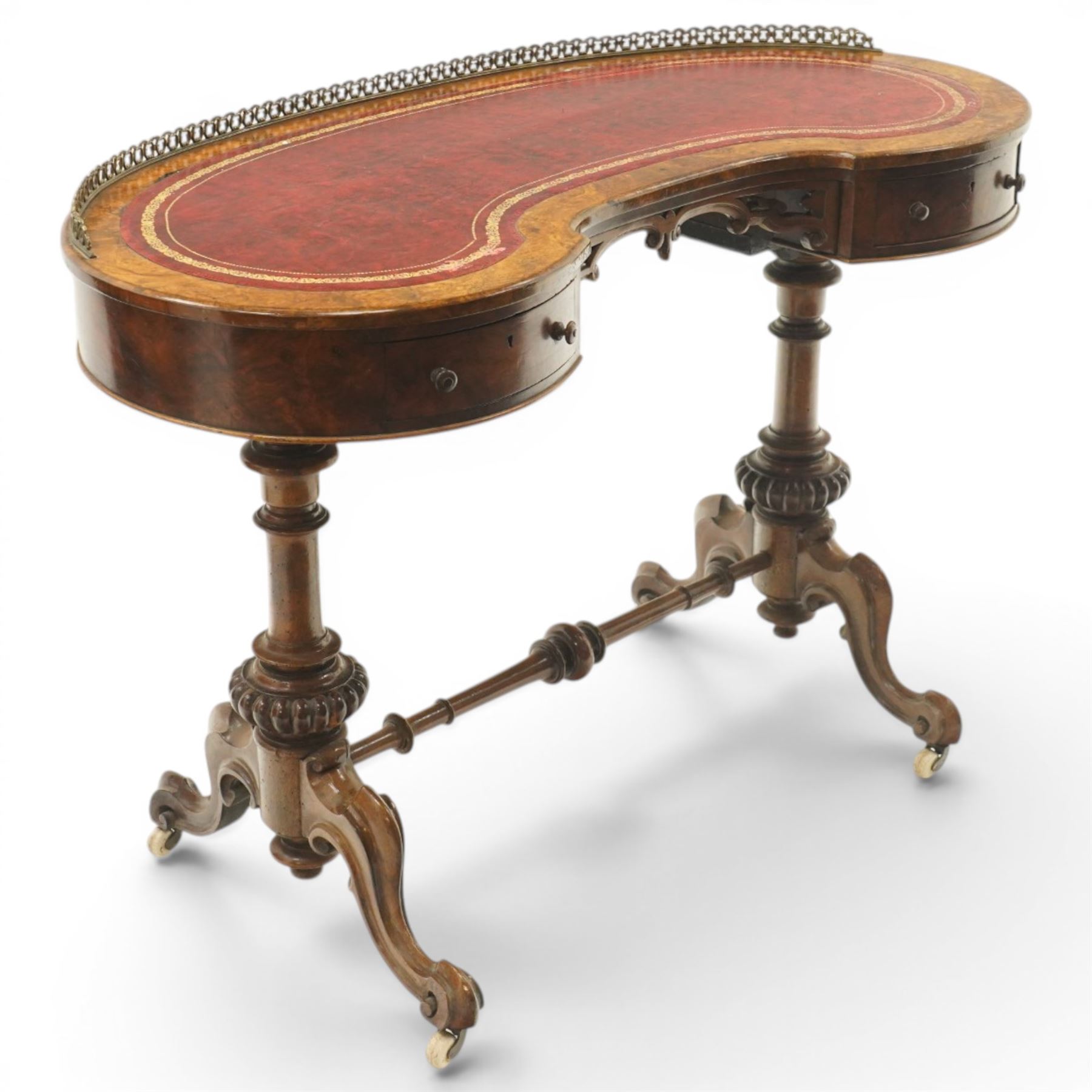 Victorian figured burr walnut kidney-shaped writing table, shaped top with pierced raised bras gallery back over inset red leather writing surface, fitted with two drawers flanking a pierced and carved scrolling apron, raised on turned and gadrooned twin pedestals with scroll-moulded cabriole supports with ceramic castors, united by ring-turned stretcher