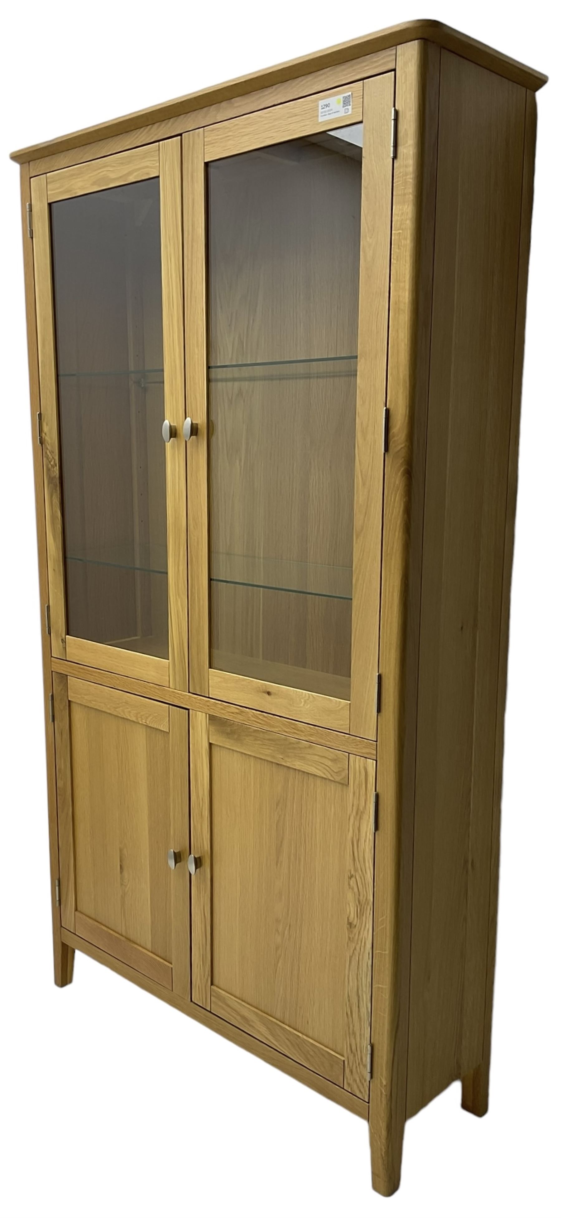 Contemporary light oak display cabinet on cupboard, fitted with two glazed doors enclosing two adjustable glass shelves with light fittings to the top, over panelled cupboard concealing single shelf, on square supports