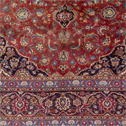 Central Persian Kashan hand-knotted carpet, madder red field with a central cusped medallion of ivory and deep indigo ground, enclosing a stylized floral rosette, framed by scrolling arabesques, field richly ornamented with an intricate network of flowering vinery, palmettes, and delicate floral sprays, spandrels in deep indigo, principal border of navy ground decorated with a continuous palmette and vine-scroll, flanked by multiple guard stripes, with cotton fringe ends 