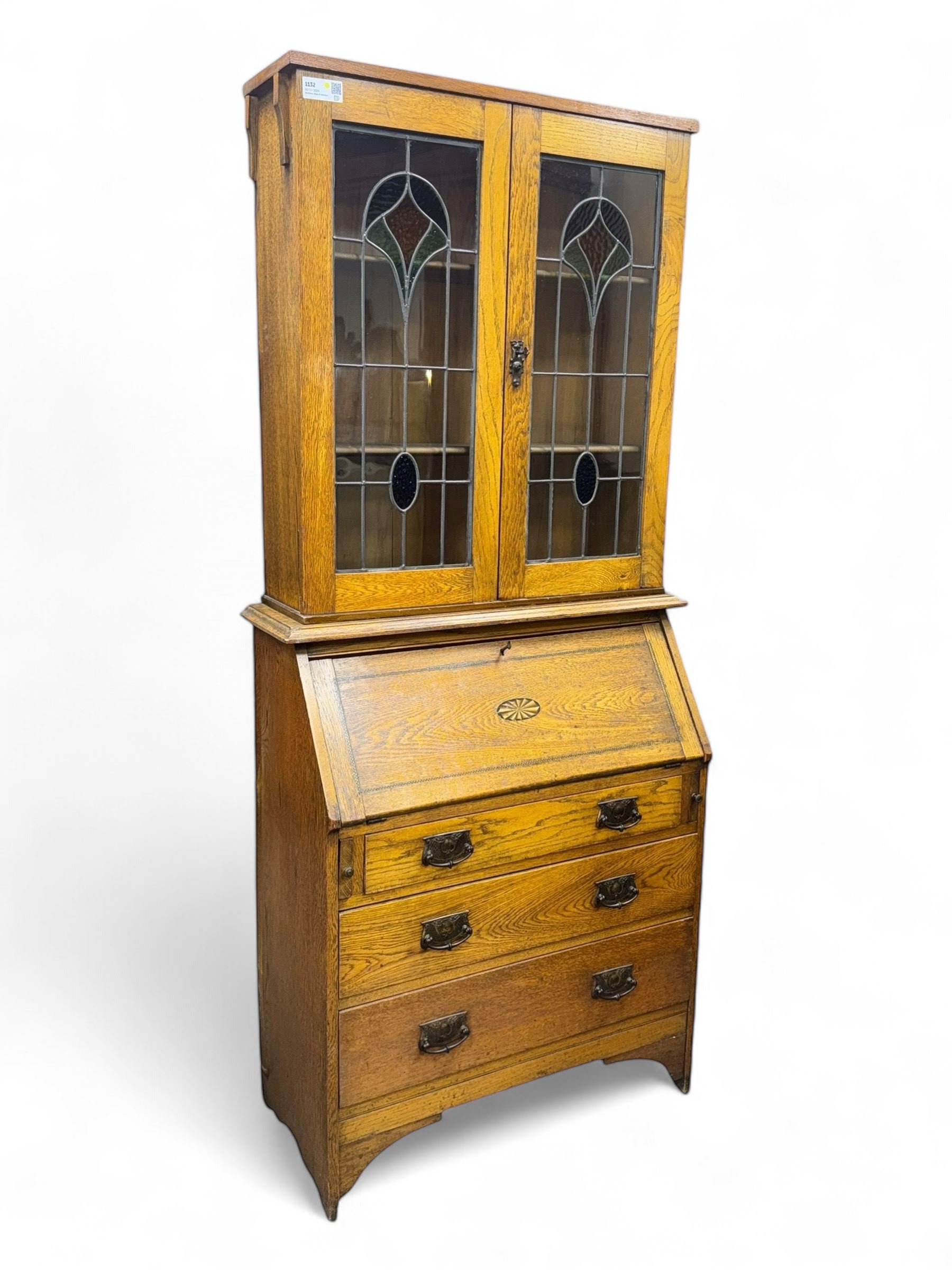 Early 20th century Arts and Crafts oak bureau bookcase, twin leaded and glazed stained glass doors enclosing two shelves, over bureau section with fall-front inlaid with sunburst motif, fitted interior of pigeonholes and shelves, on bracket feet