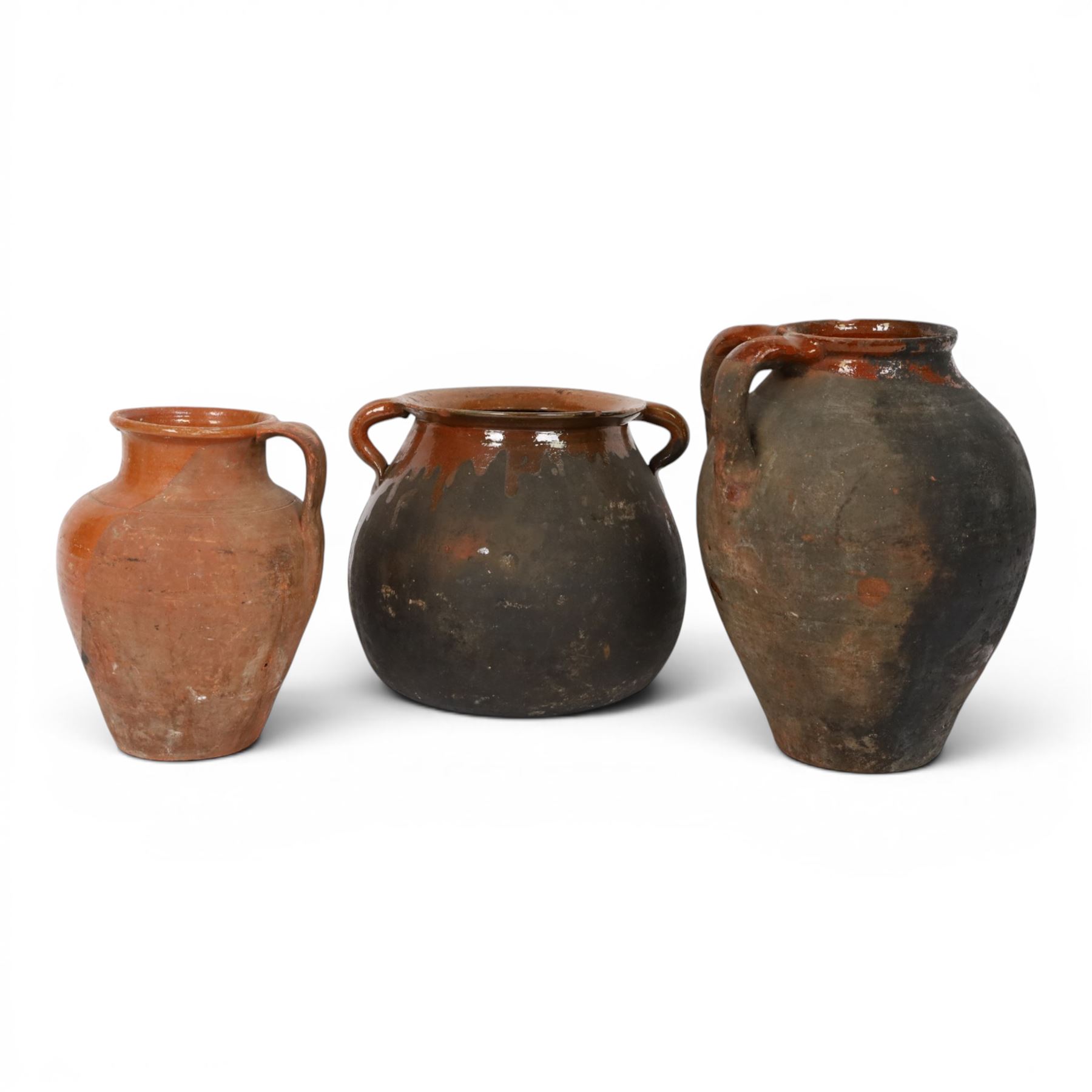 Three early 20th century Spanish part glazed terracotta storage vessels, the first example of bellied form with rounded base and twin loop handles, H24cm, the second of baluster for with close set loop handles, H30cm, and the third smaller also of baluster form with single loop handle, H24cm