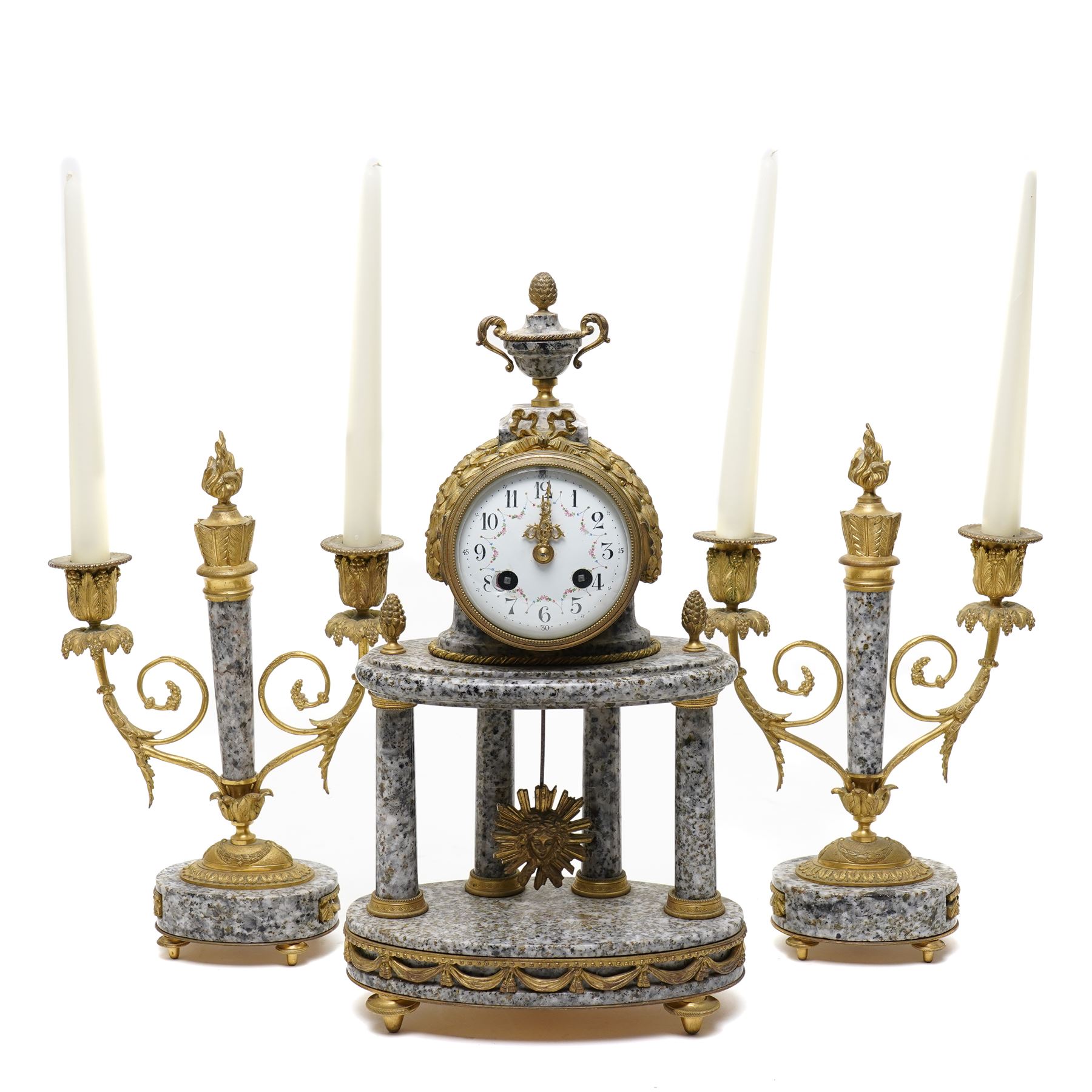 French- early 20th century portico clock garniture, 8-day drum movement decorated with laurel leaves to the glazed bezel and surmounted by a decorative urn with scroll handles, raised by four matching columns on a granite effect oval base with toupee feet, convex enamel dial with gilt pierced hands, Arabic numerals, minute markers and a garland of flowers, twin train Parisian rack striking movement, striking the hours and half hours on a bell. With a sunburst pendulum and key and a matching pair of two-light candelabra.