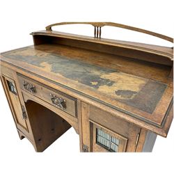 Arts and Crafts period twin pedestal desk, arching pediment on spindle supports, raised shelf on moulded rectangular top with leather inset, fitted with central drawer and two cupboards, the doors with lead glazed panes, pressed copper handle plates 
