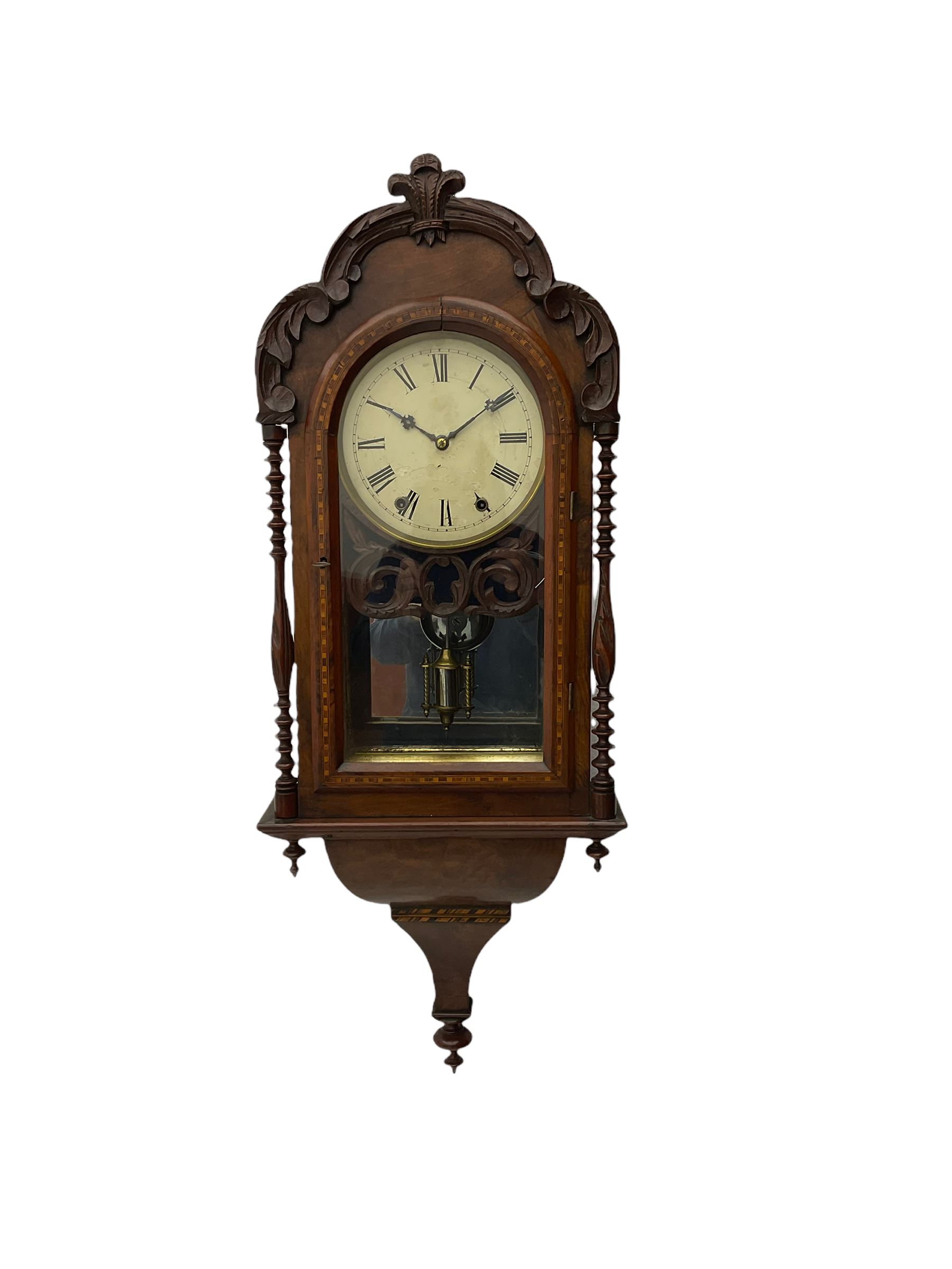 A 19thcentury American wall clock in a walnut case with crossbanding, with a carved pediment surround and arched glazed door flanked by two ring turned columns with pendants on an ogee plinth beneath, with a spring driven movement striking the hours on a bell, eight-inch painted dial with roman numerals, minute track and steel Maltese-Cross hands, with a spun brass bezel and winding collets, visible mock mercury pendulum with a reflective silvered mirror to the rear. With key and pendulum.   

