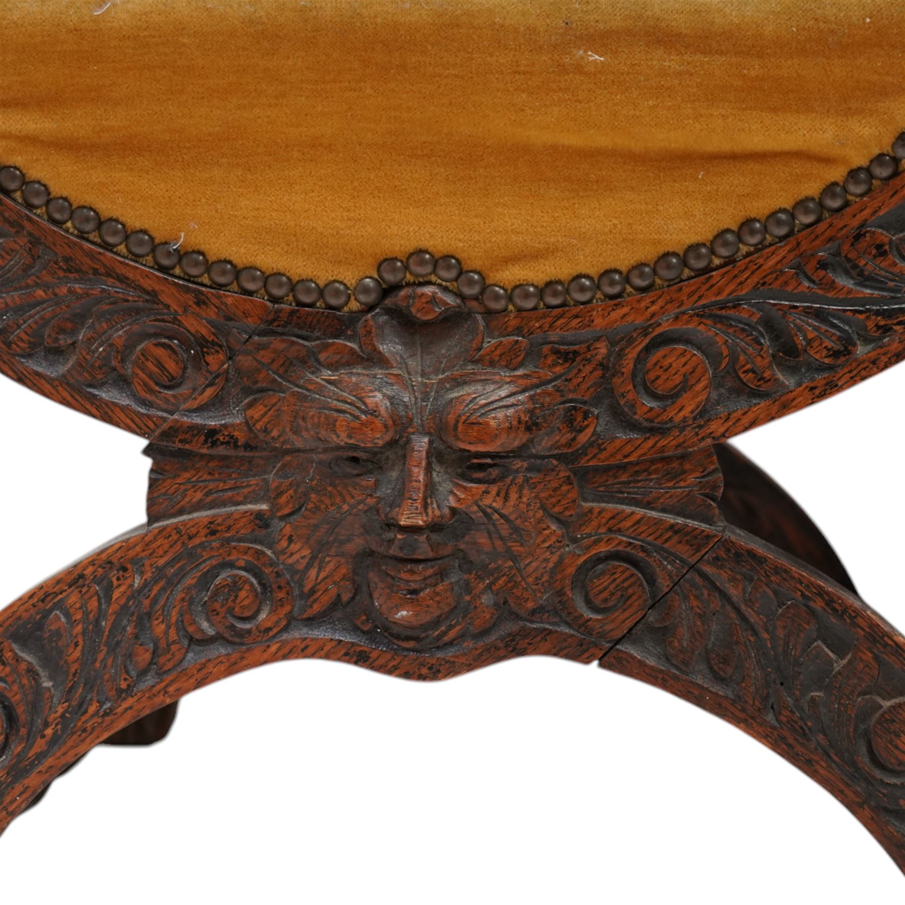 Victorian heavily carved oak X-framed throne chair, the cresting rail carved with mask and curling foliage, upholstered in mustard fabric, the arms and frame carved with scrolling foliage, united by turned stretchers 