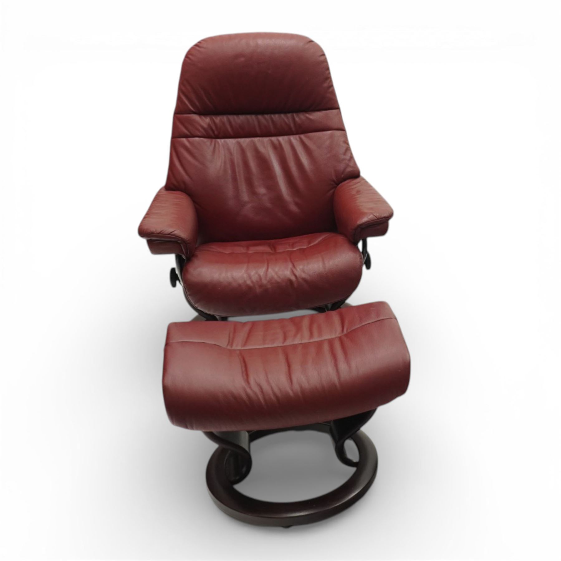 Ekornes - Stressless reclining armchair and matching footstool, upholstered in oxblood leather with padded headrest and segmented back cushion, shaped armrests with exposed adjustment handles, raised on stained laminated beech circular swivel base, the conforming stool on matching ring base