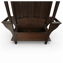 Arts and Crafts design oak settle, with projecting moulded cornice over a boarded back fitted with a rectangular bevelled mirror plate and a row of wrought iron coat hooks, the panelled back with arched fielded panels, flanked by shaped umbrella or stick stands with removable drip trays, the plank hinged seat over a panelled box base, on square section supports