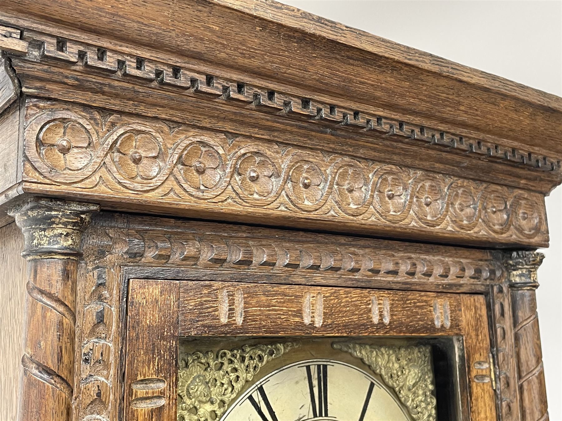 19th century carved oak longcase clock, projecting cornice over guilloche carved frieze, square brass dial with silvered Roman chapter ring and calendar aperture, decorated with ornate mask cast spandrels, the trunk door and base carved with potted plant, twin train driven eight day movement striking on bell
