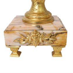 Pair of Rococo style gilt metal table lamps, the urn shaped bodies each with applied figure seated upon shoulder, and female mask to opposite side issuing scroll leading to top of neck, the body cast and applied with stiff leaf detail, and foliate swags and wreaths, upon square pink marble plinths with applied gilt metal mask mounts, and four gilt metal bracket feet, each supported a pleated cream silk closed top lampshade, H63cm