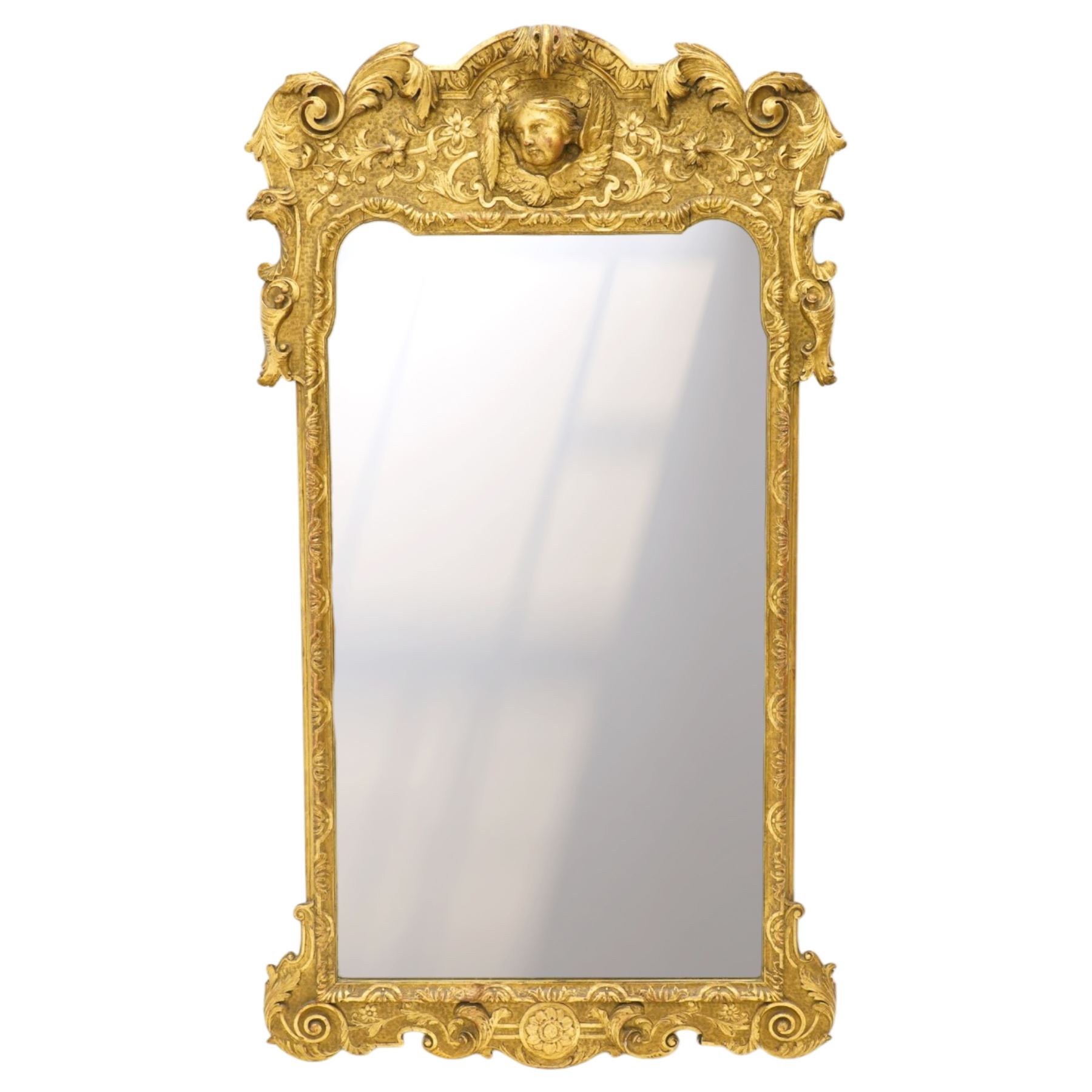 In the manner of Thomas Johnson - circa 1900 Georgian Revival giltwood wall mirror, the shaped rectangular plate within a foliate and scallop carved slip and richly ornamented frame, surmounted by a pierced cartouche crest centred with a protruding relief carved winged cherub mask and issuing scrolling acanthus and floral ornament, the sides with eagle masks over acanthus scrolls, terminating in a shaped apron with moulded rosette and spiralling acanthus leaves