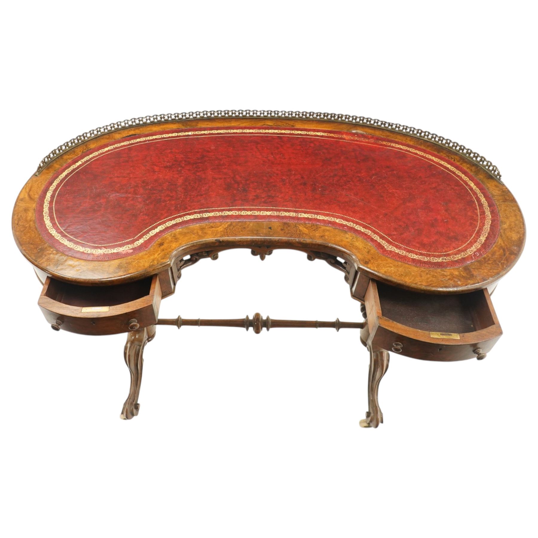 Victorian figured burr walnut kidney-shaped writing table, shaped top with pierced raised bras gallery back over inset red leather writing surface, fitted with two drawers flanking a pierced and carved scrolling apron, raised on turned and gadrooned twin pedestals with scroll-moulded cabriole supports with ceramic castors, united by ring-turned stretcher