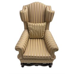 William and Mary style wingback armchair, upholstered in gold striped fabric, on walnut scroll carved supports joined by scroll and foliate carved middle rail, turned H-shaped stretchers, on castors