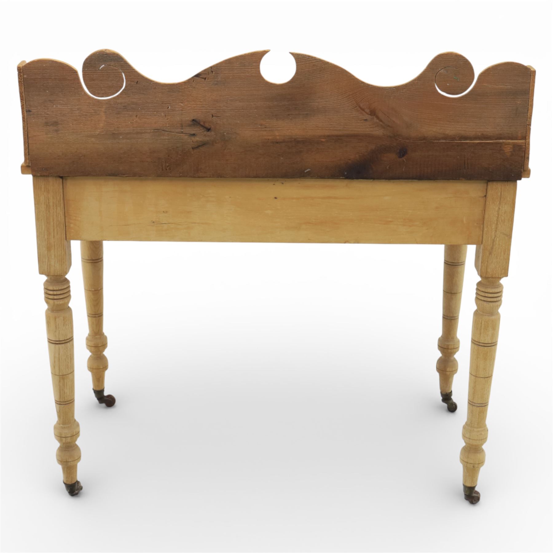 19th century stripped pine side table, the rectangular plank top with low shaped back rail incorporating scroll cut ends, fitted with two frieze drawers with ceramic knob handles, raised on turned supports terminating in castors